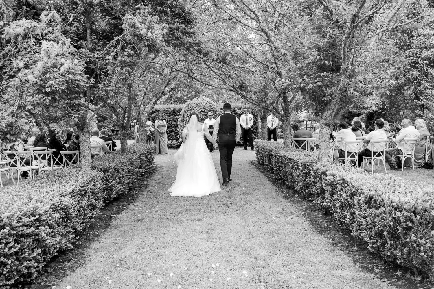 Black and white photo of Janine walking down a hedge-lined aisle