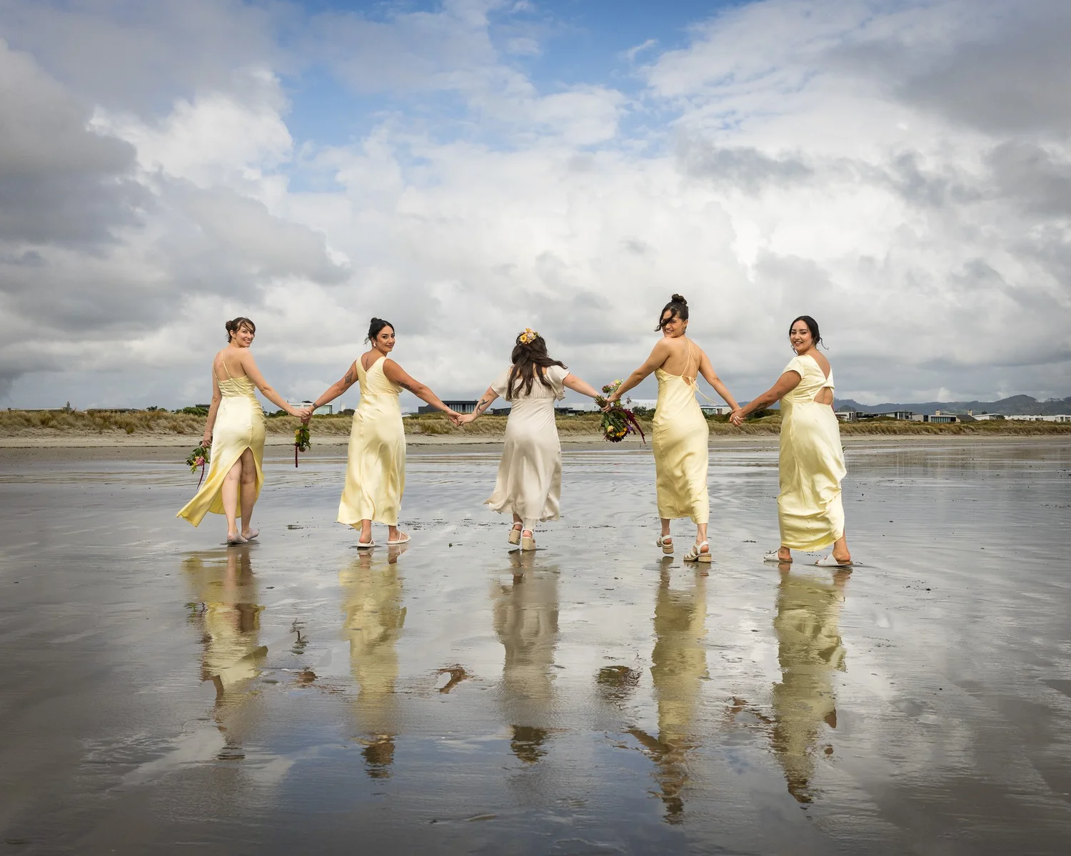 The bridesmaids walk hand in hand across the wet sand