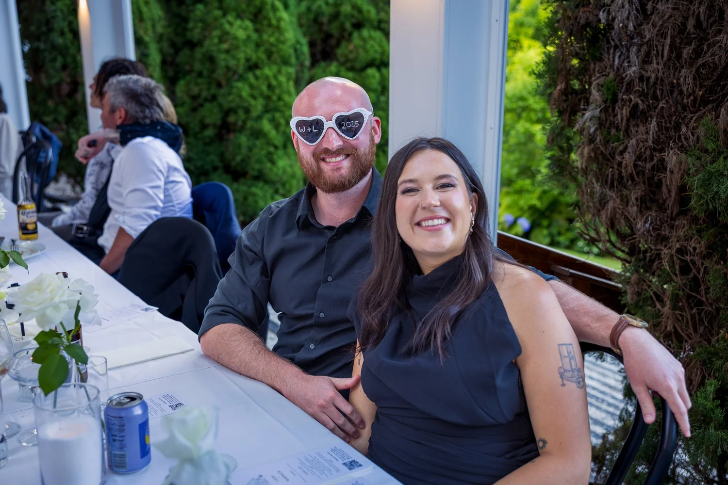 A guest in heart-shaped sunglasses smiles beside his partner