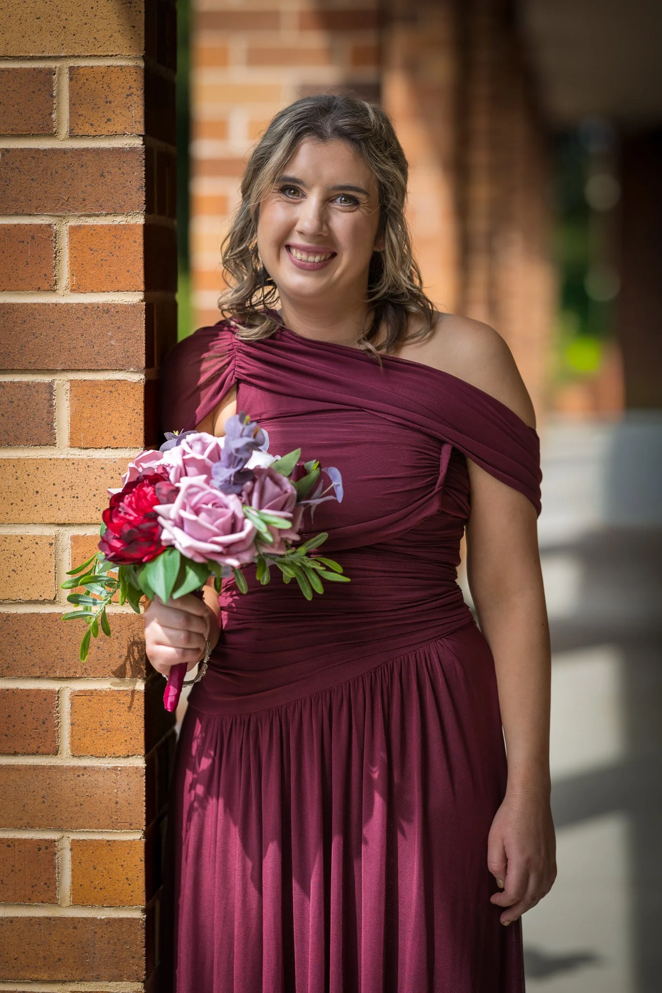 A bridesmaid in burgundy holds her bouquet beside a brick column