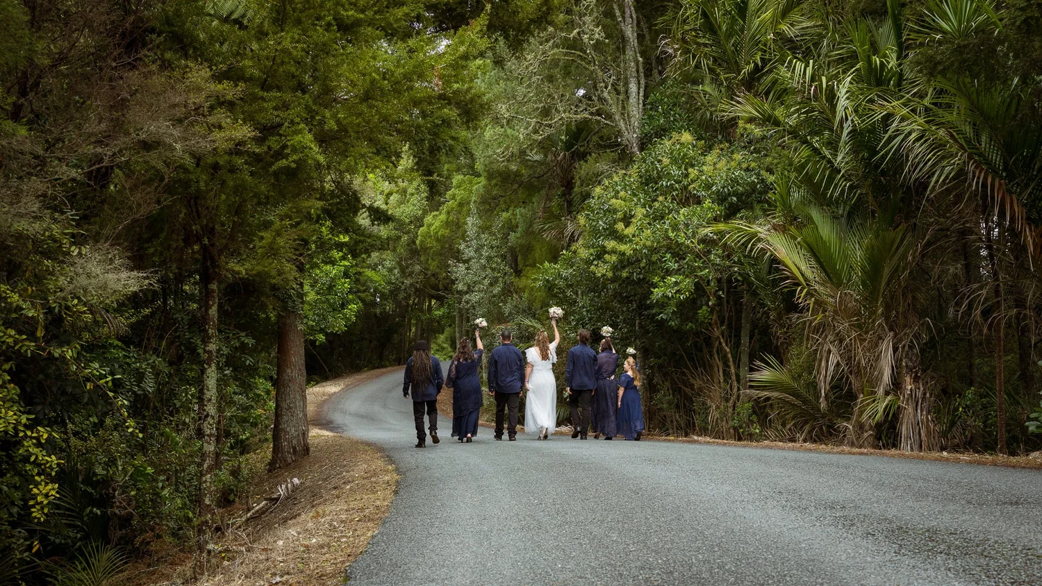 The wedding party walks down a bush-lined road