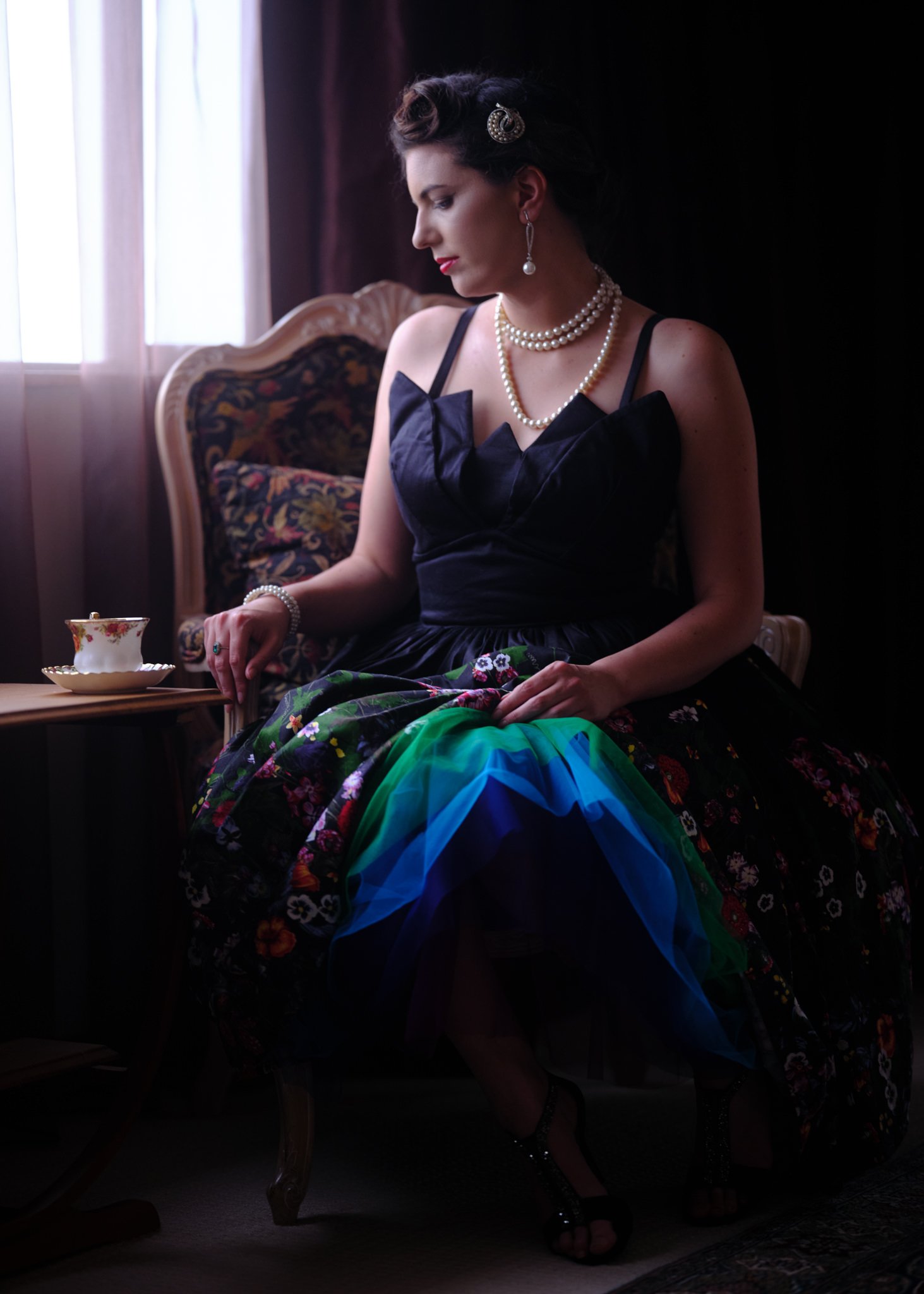 A woman in pearls and a colourful skirt taking tea by a window