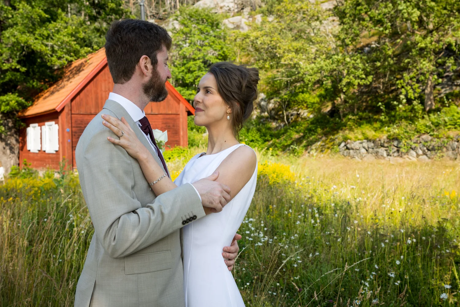 Sofia and John embrace in a meadow beside a red cottage