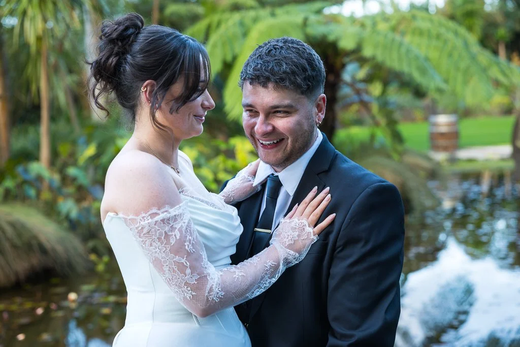 A bride smiles at her groom beside a garden pond