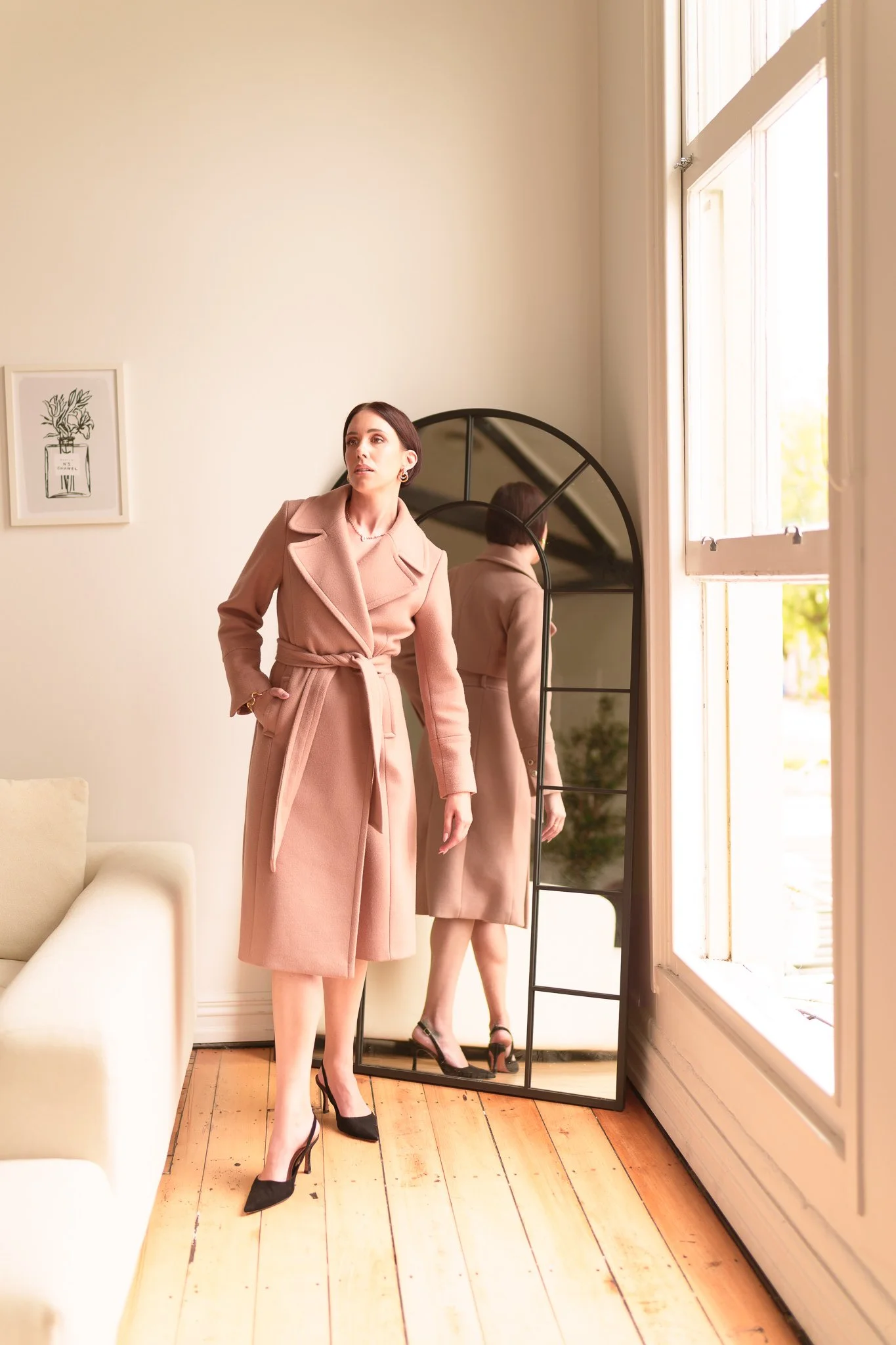 A woman in a blush coat standing by a tall window
