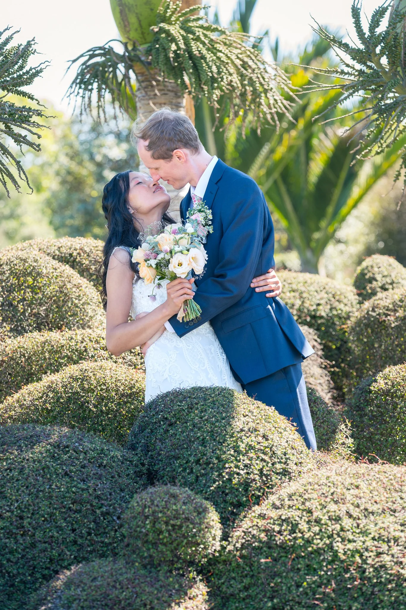 A bride and groom kiss among clipped topiary in a garden