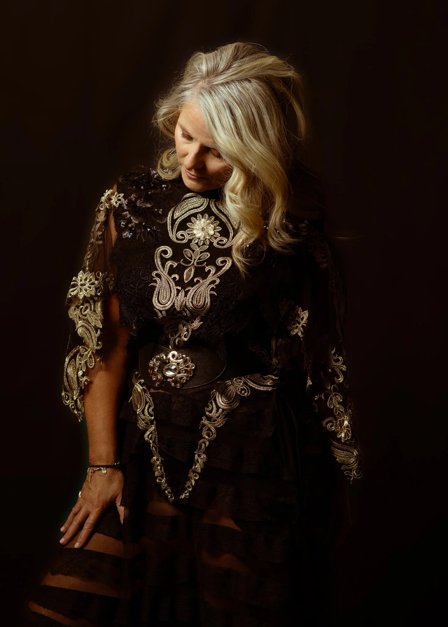 A woman looks down in an embroidered black gown in low light