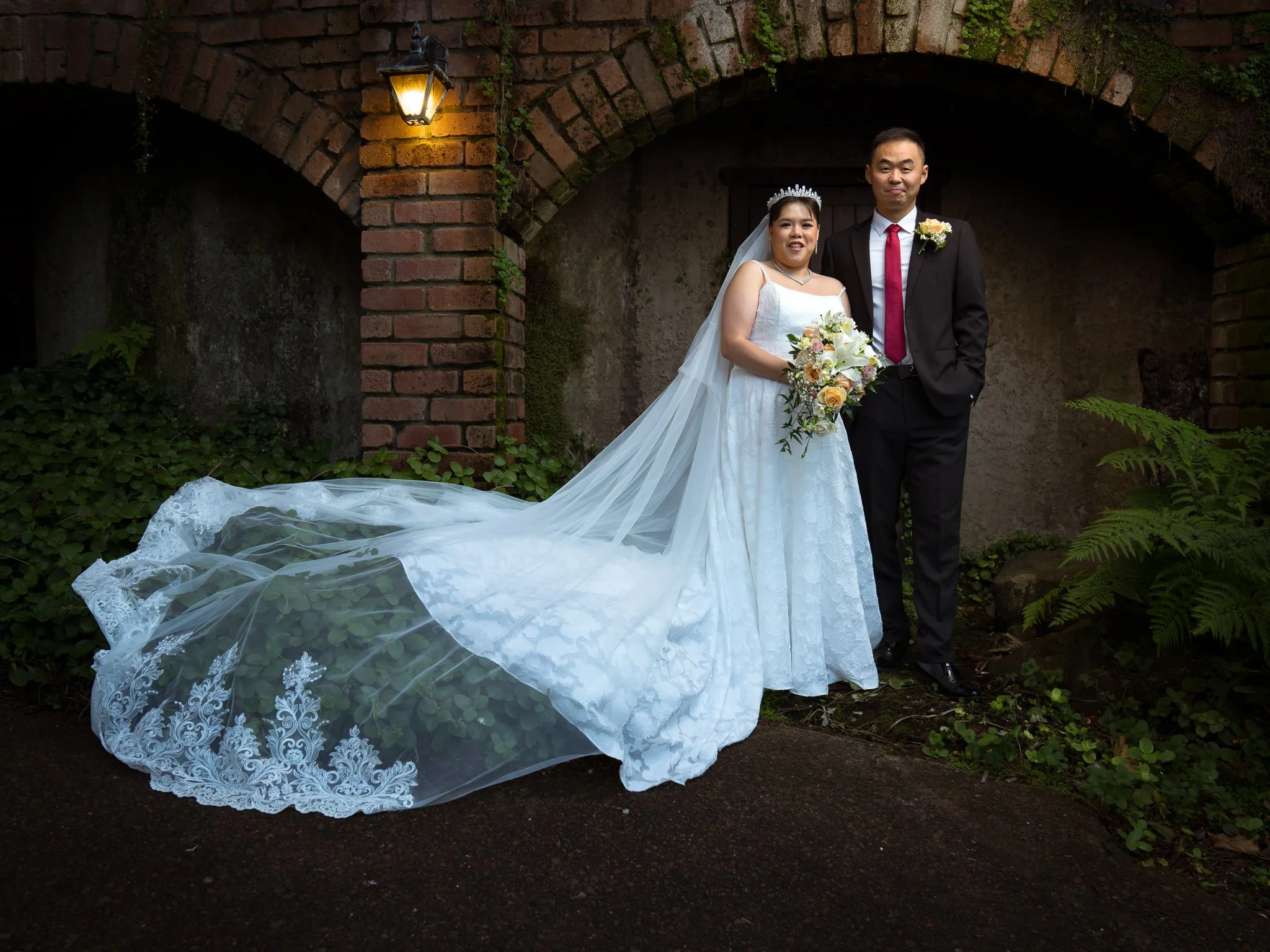 A bride and groom standing together outdoors under an arched brick structure, dressed in wedding attire, with the bride holding a bouquet of flowers and the bride wearing a tiara and a long veil.