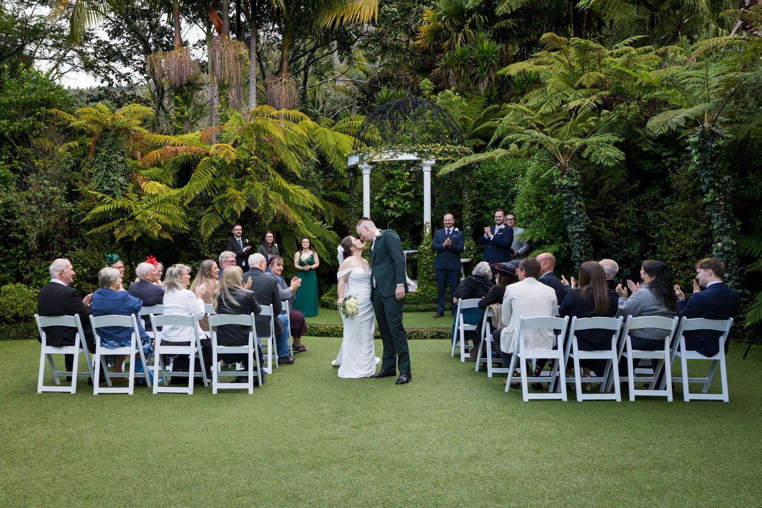 A couple stand before guests under a white arbour in the bush