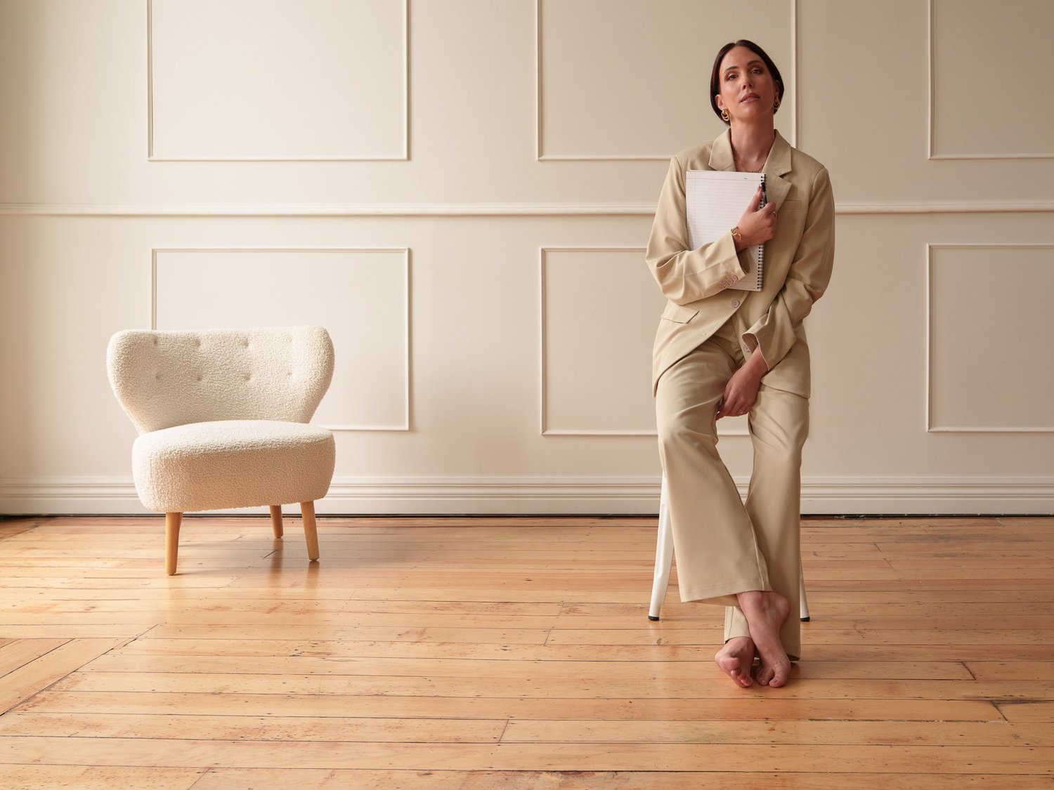 A woman in a beige suit perched on a stool in a panelled room