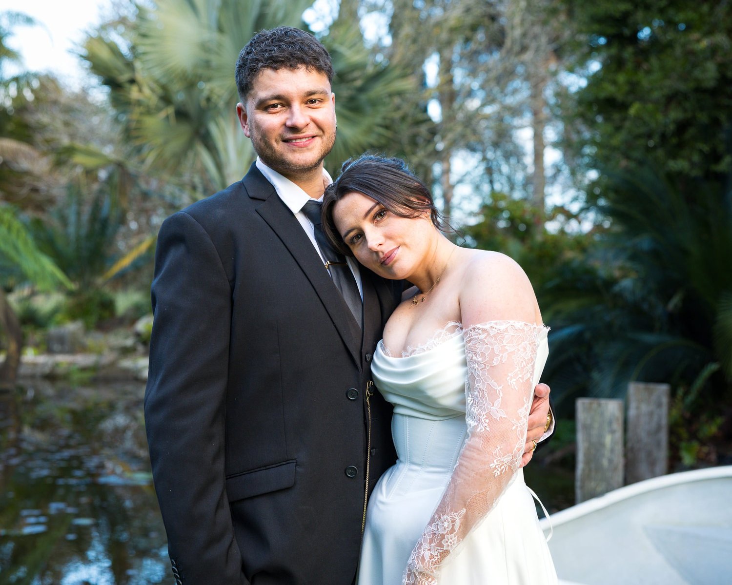 A couple pose beside a pond and rowing boat