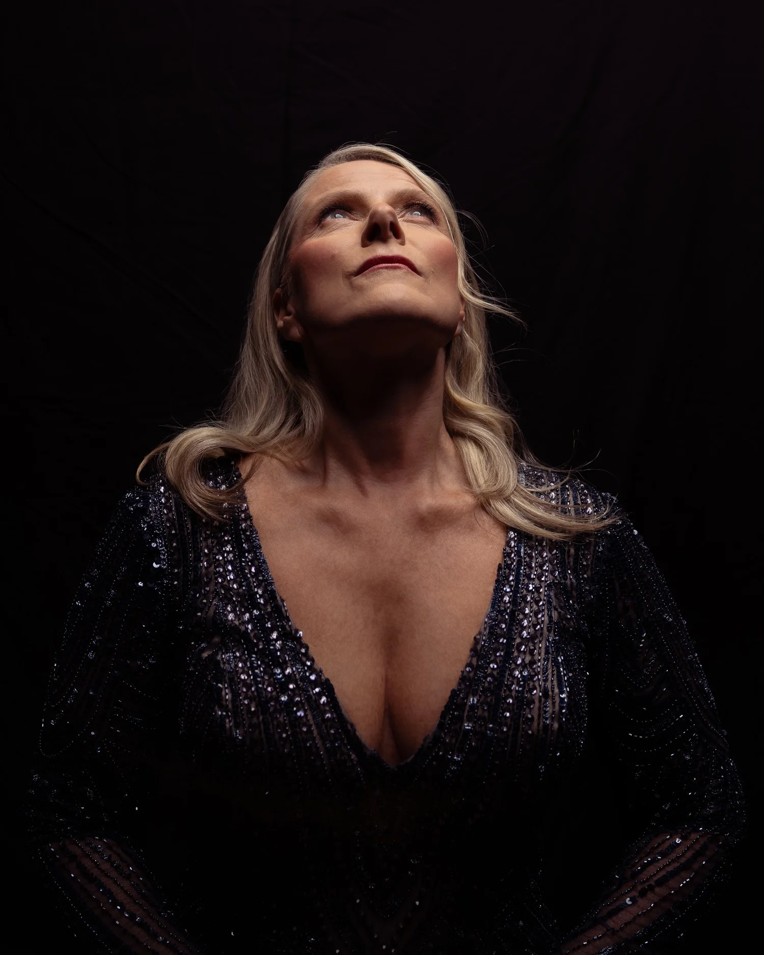 A woman in a sparkling gown looks upwards in dramatic light