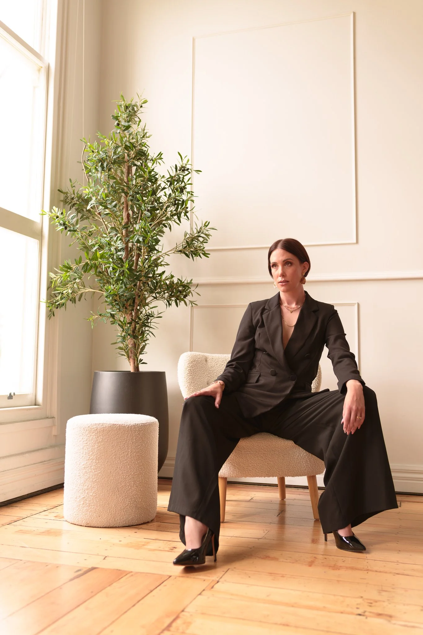 A woman in a black suit seated beside a potted olive tree