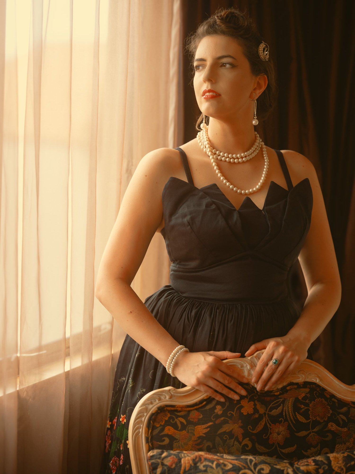 A woman in a black dress and pearls seated by a window