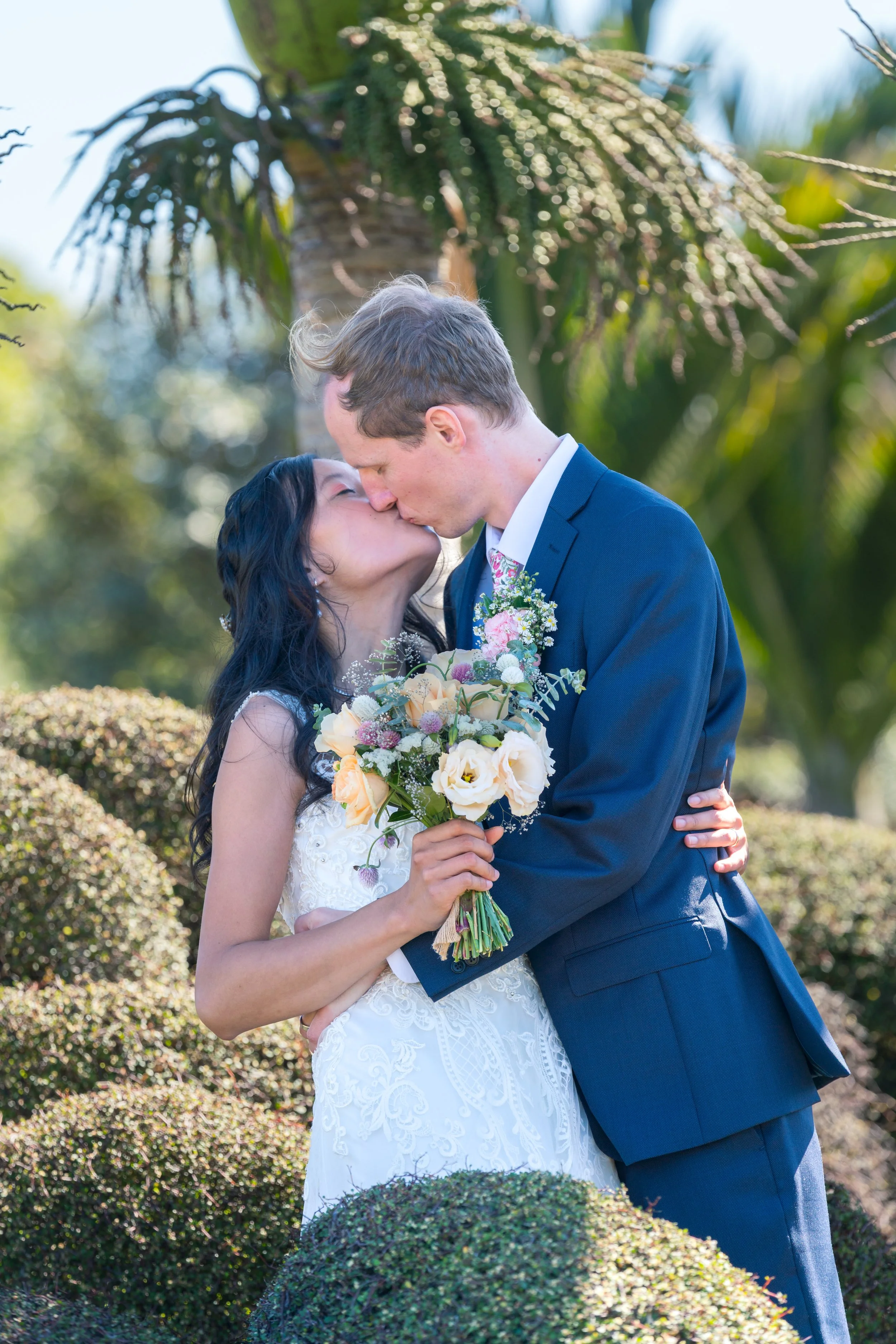 A bride and groom share a kiss outdoors, with green bushes and trees in the background, the bride holding a bouquet of flowers.