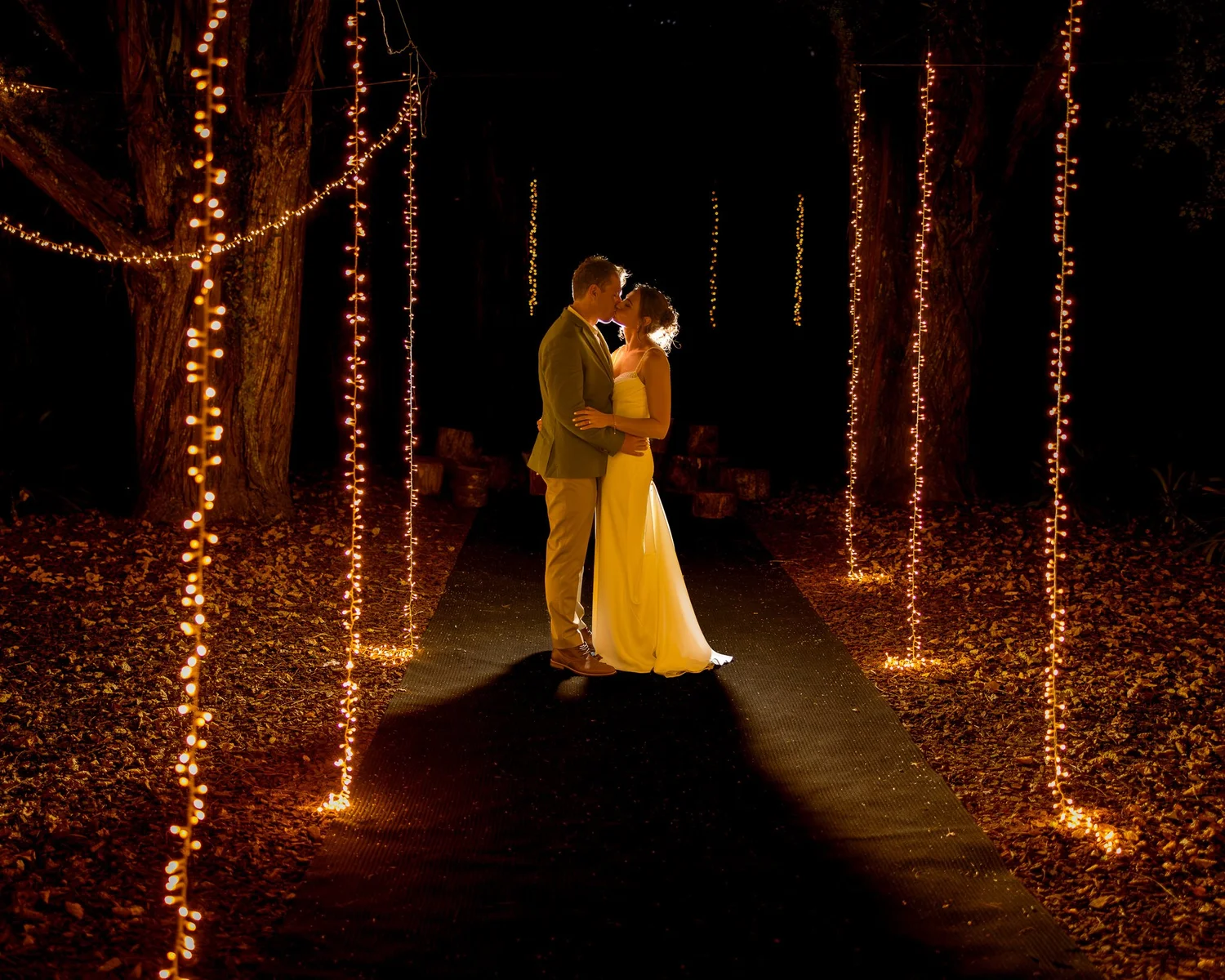 Jade and Ben embrace between trees strung with fairy lights at night
