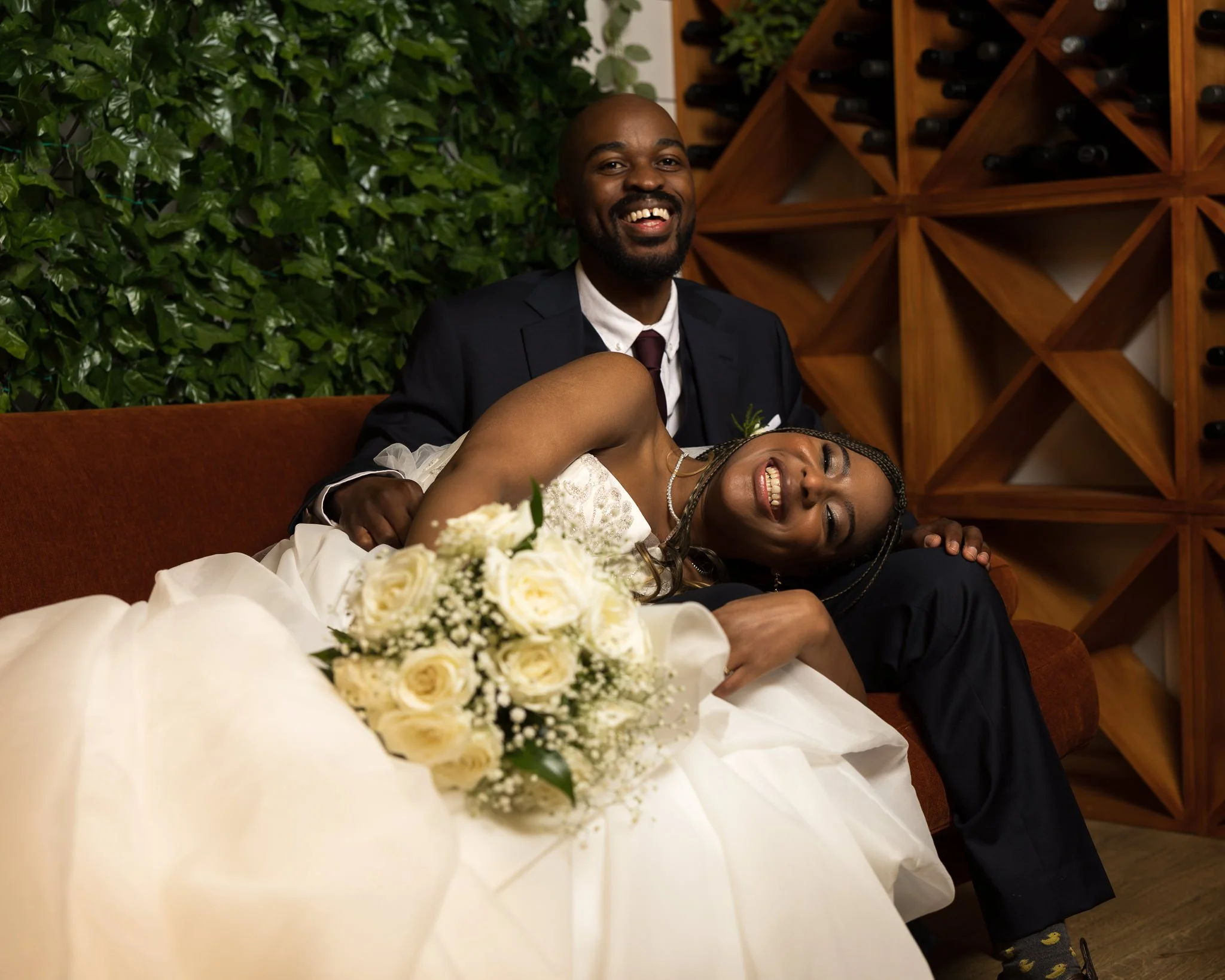 A smiling couple on a brown couch, the woman in a wedding dress holding a bouquet of white roses, and the man in a tuxedo, laughing with their heads close together, in front of green foliage and a wooden wine rack.