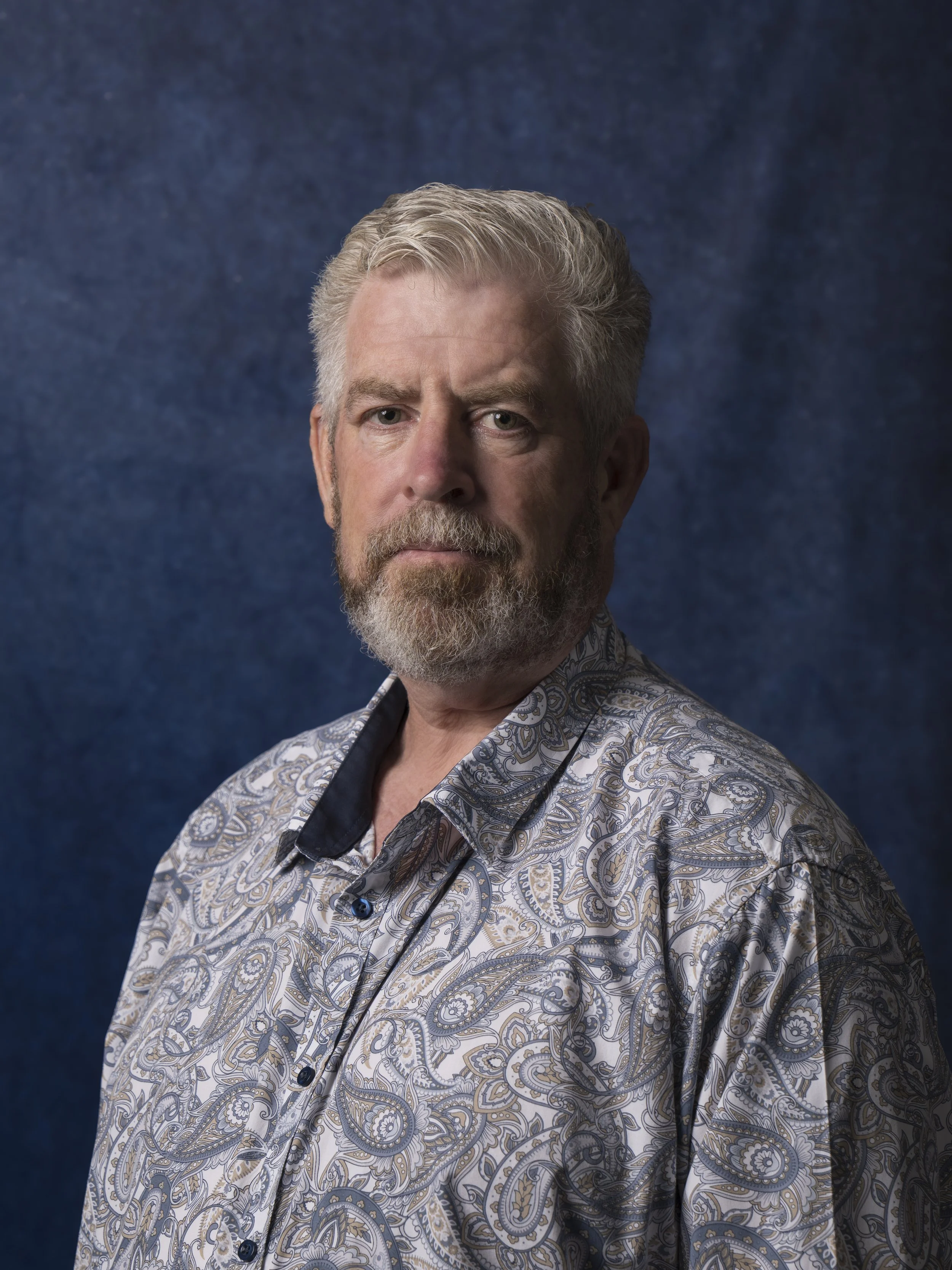 Portrait of a middle-aged man with gray hair and beard, wearing a paisley patterned shirt, against a textured blue background.
