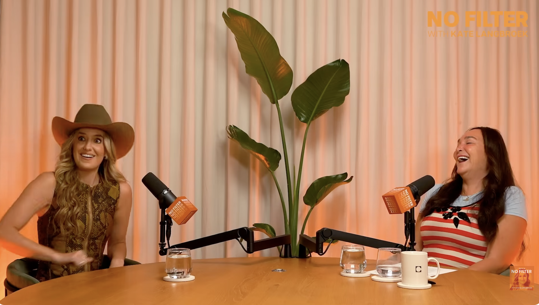 Two women are in a podcast studio with orange microphones that say 'No Filter with Kate Langbroek.' They are sitting at a round wooden table with two glasses of water and a white mug. A large plant with broad green leaves is between them, and the background has vertical cream-colored curtains.