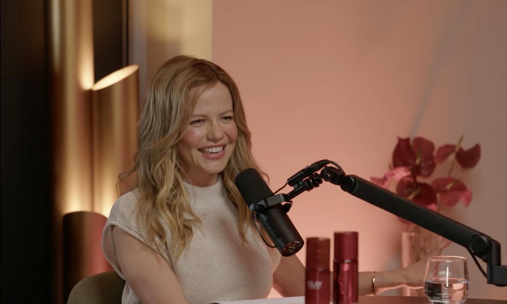 Woman with blonde wavy hair smiling while speaking into a microphone during a recording or podcast session, with bottles and a glass of water on the table, in a warmly lit room.