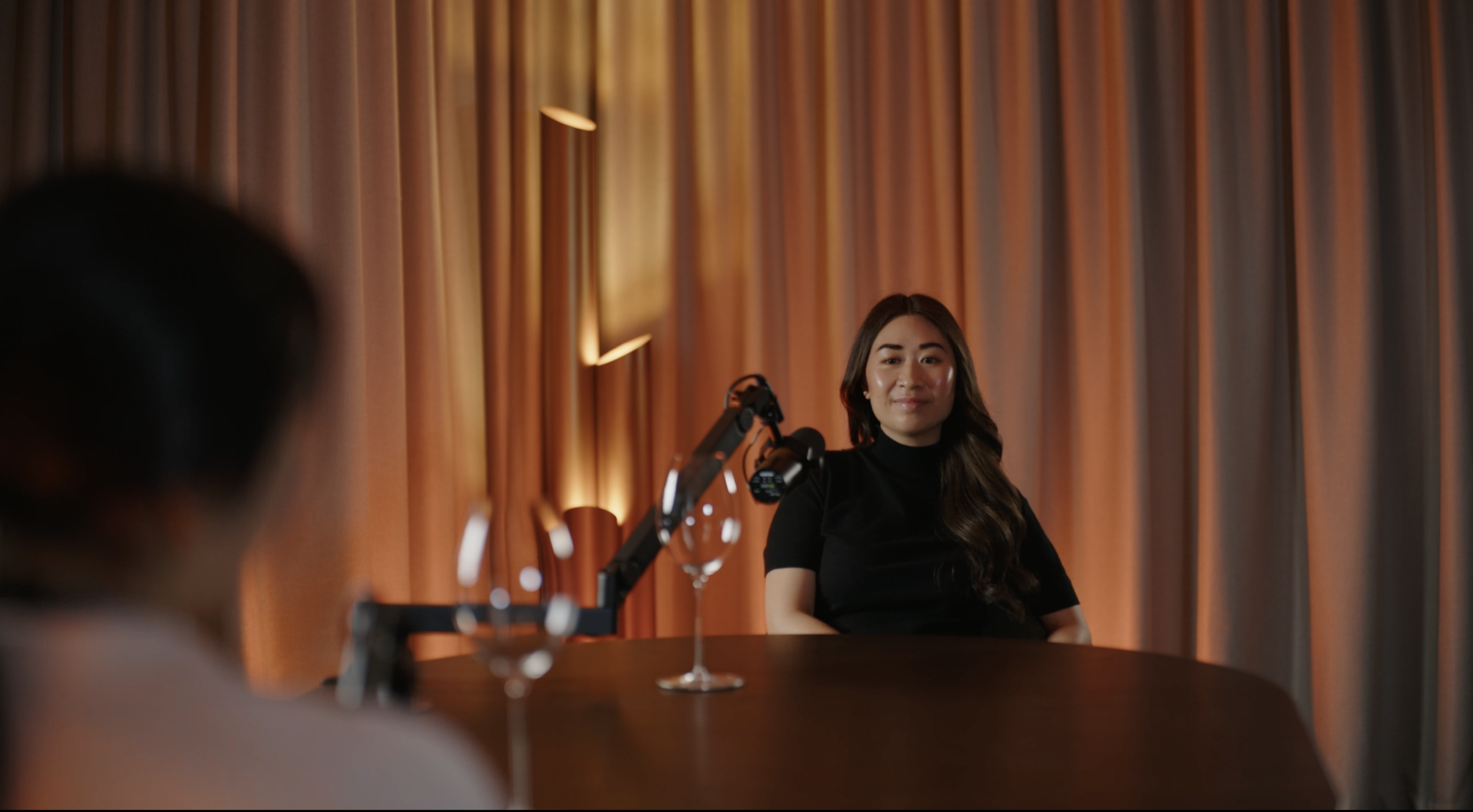 A woman in a black dress sitting at a table with a microphone and glass of water, facing another person, against a backdrop of orange curtains and warm lighting.