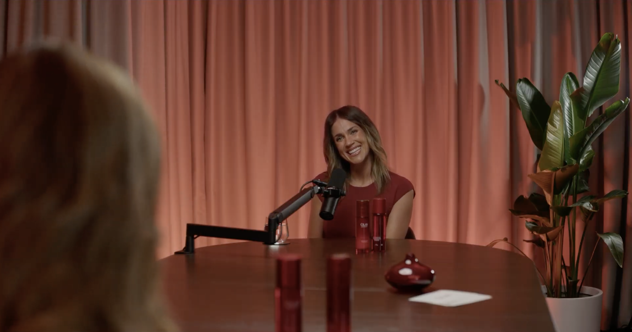A woman with shoulder-length brown hair smiling during an interview or podcast, seated at a table with a microphone, in front of an orange curtain, with green potted plant to the right.