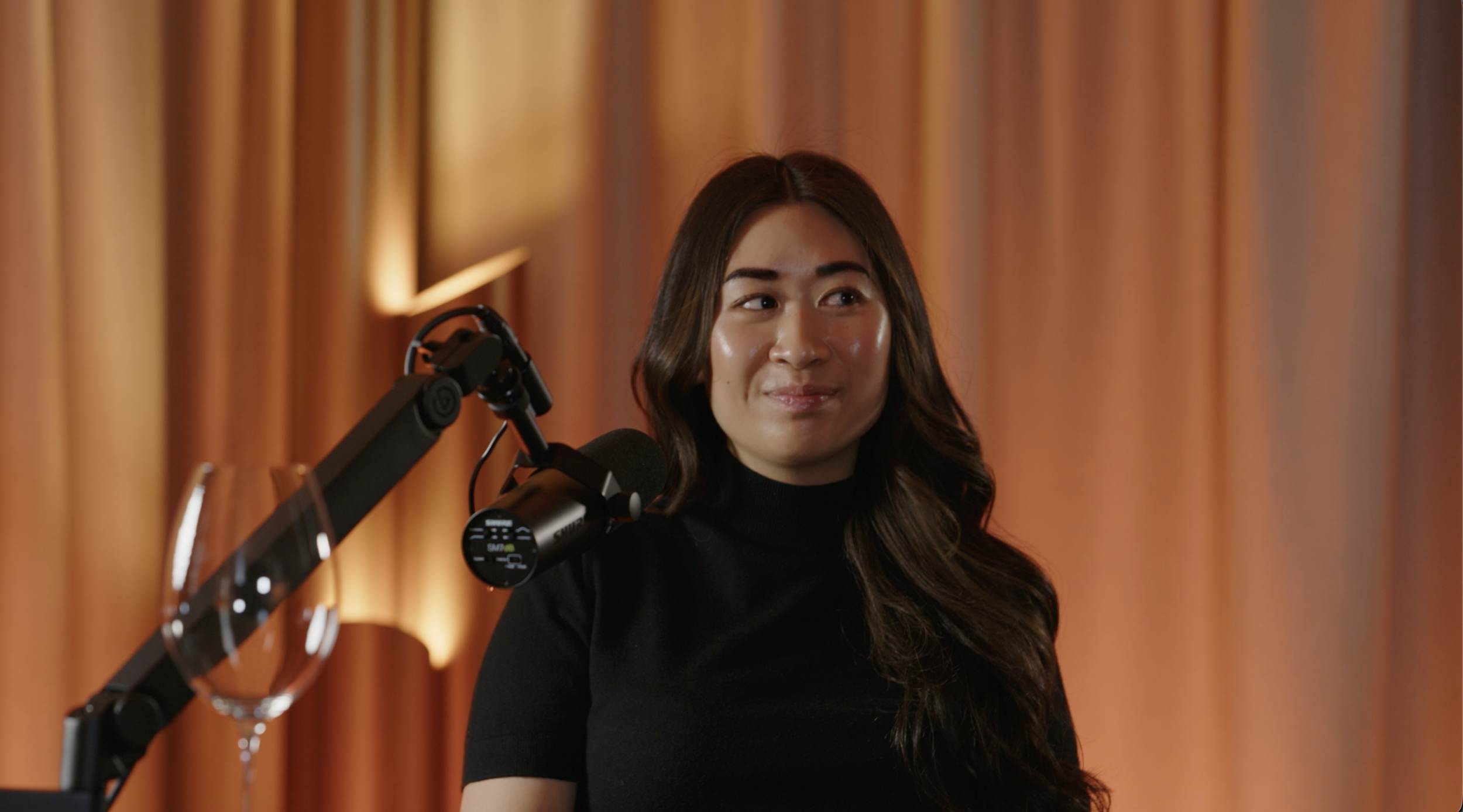 A woman with long dark hair wearing a black turtleneck, sitting at a podcast or radio studio with orange curtains in the background, next to a microphone and a wine glass.