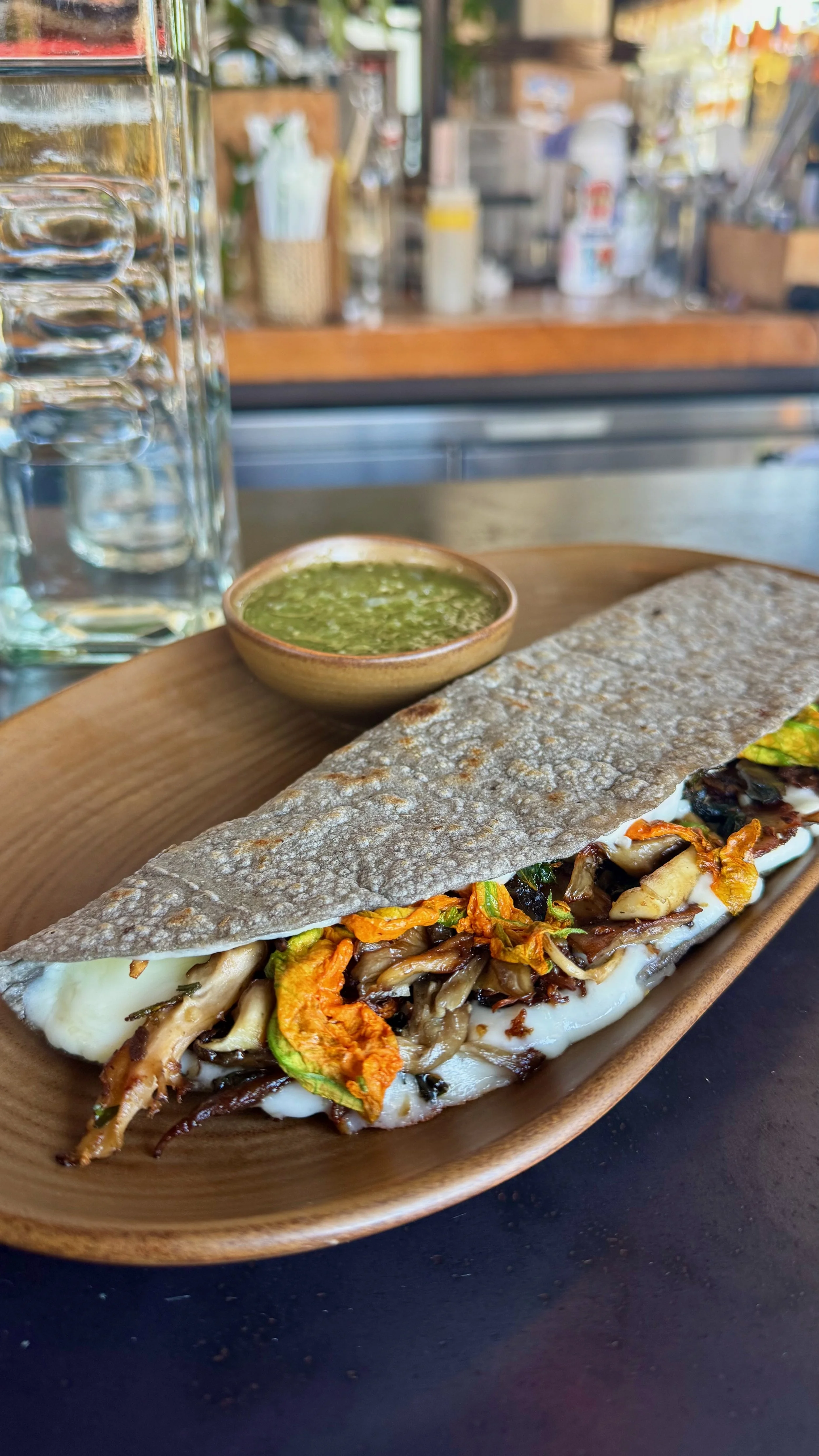 A veggie torta sandwich on a wooden plate with a side of green sauce in a small bowl, next to a glass of water, on a black surface in a casual dining setting.
