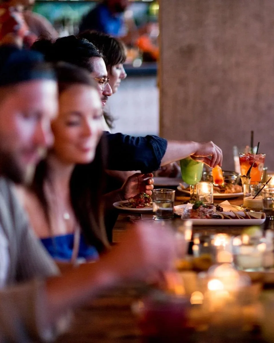 People sitting at a restaurant table with drinks and food, some of them appear to be engaged in conversation or serving themselves beverages, with dim lighting and a cozy atmosphere.