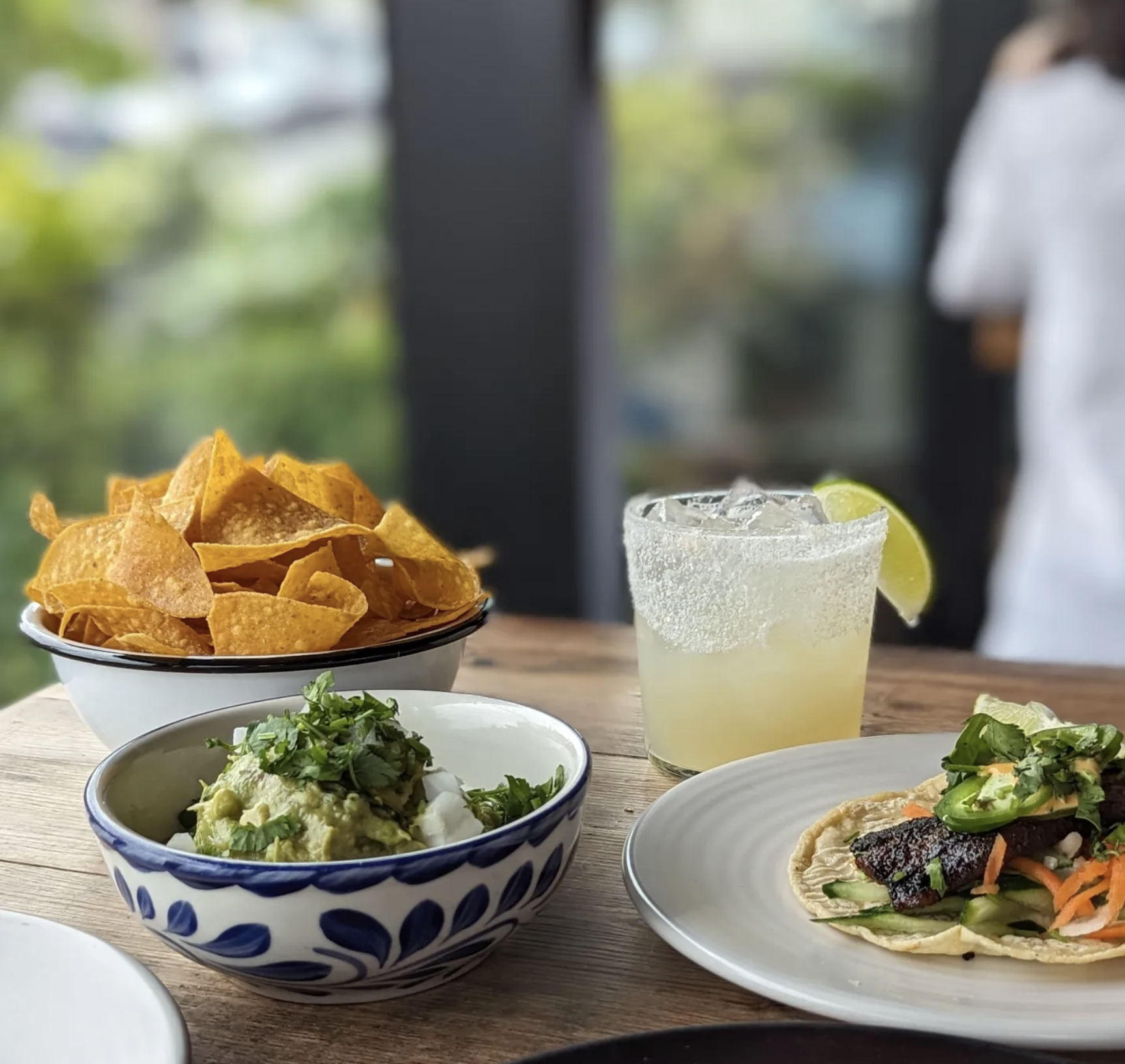 A table set with a bowl of tortilla chips, a bowl of guacamole, a plate of tacos with fish and vegetables, and a glass of lime margarita with salt on the rim. In the background, there are blurred plants and a window.