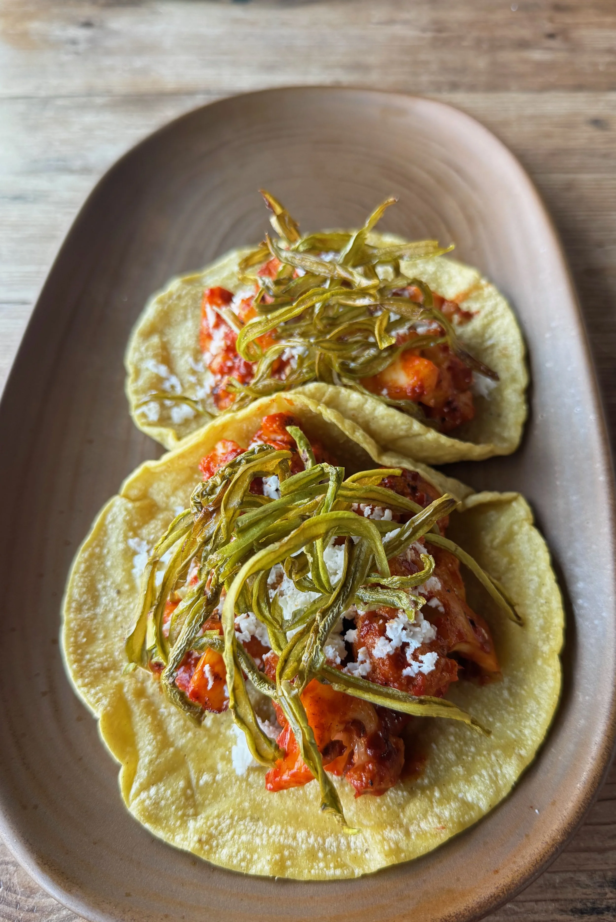 Three tacos with yellow corn tortillas topped with shredded cheese, green peppers, tomato sauce, and diced vegetables, served on a beige oval plate.