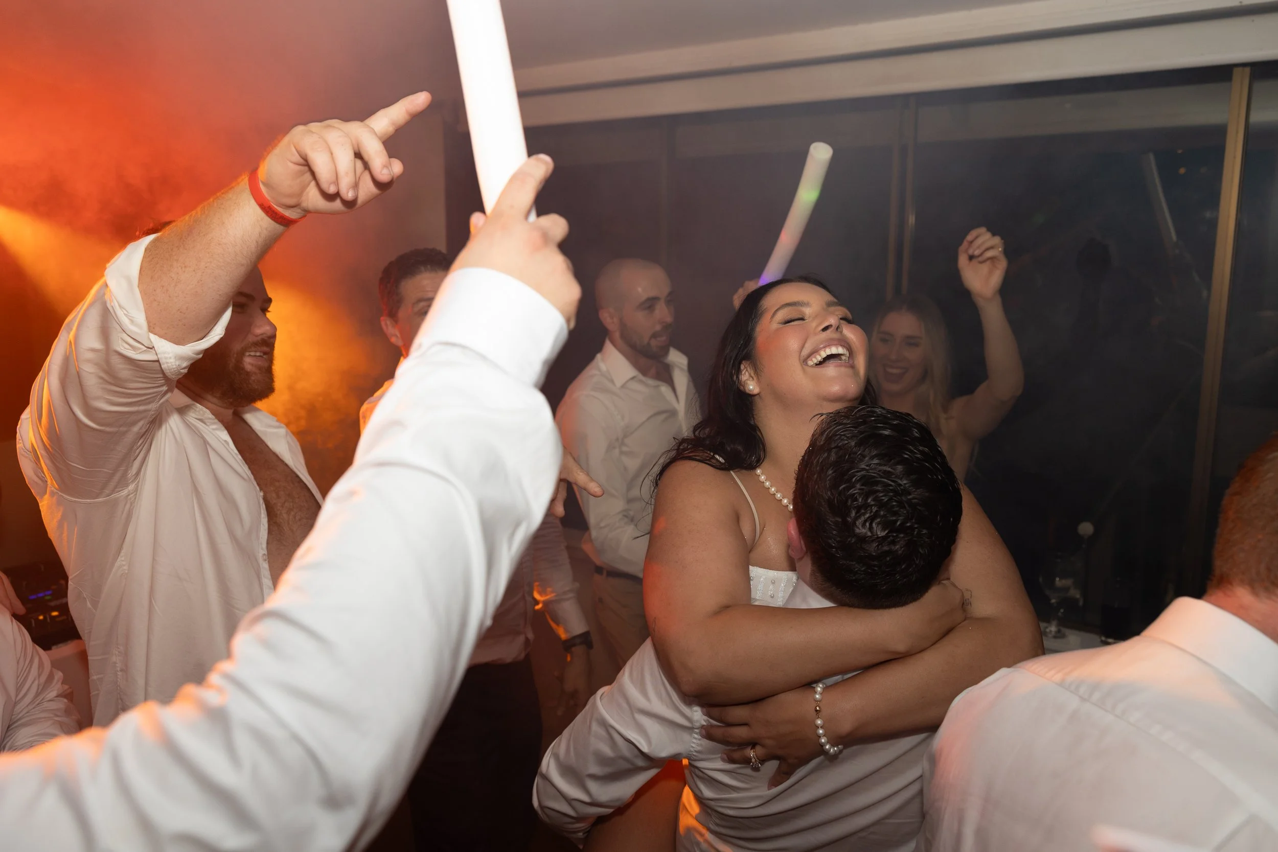 Group of people celebrating, with a woman holding a man in a dance embrace, smiling and enjoying the moment at a lively party or wedding reception.