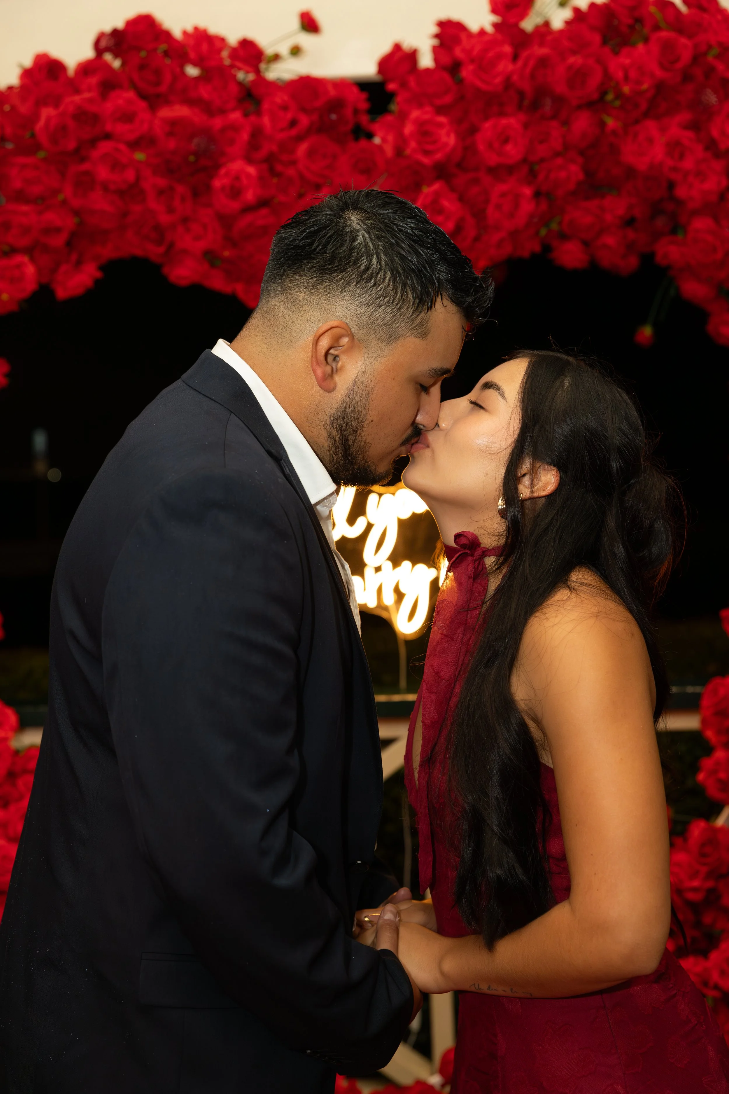 A couple sharing a kiss at a romantic event, with flowers and a neon sign in the background.