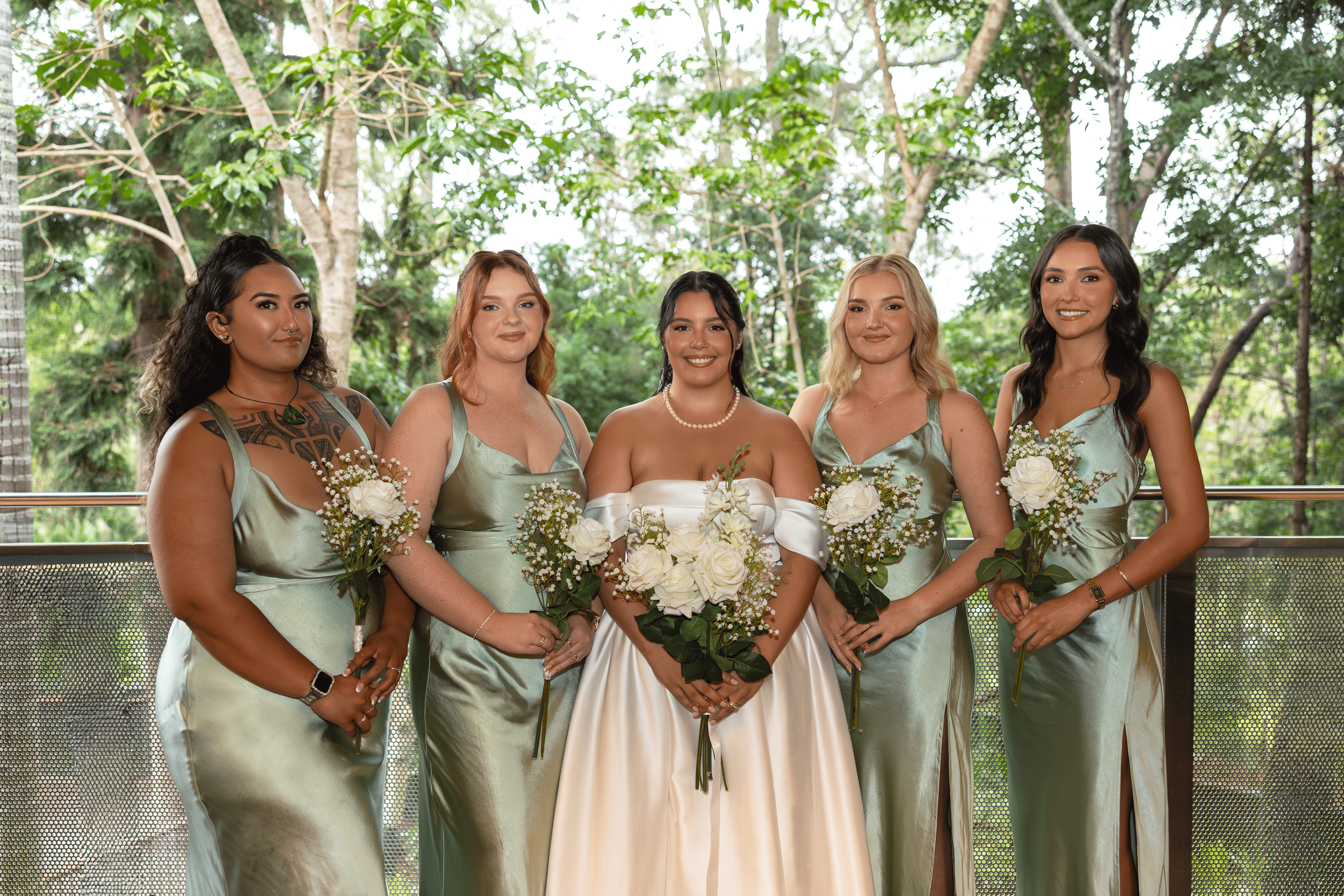 Bride and four bridesmaids standing together outdoors at a wedding, all holding bouquets of white flowers, with lush green trees in the background.