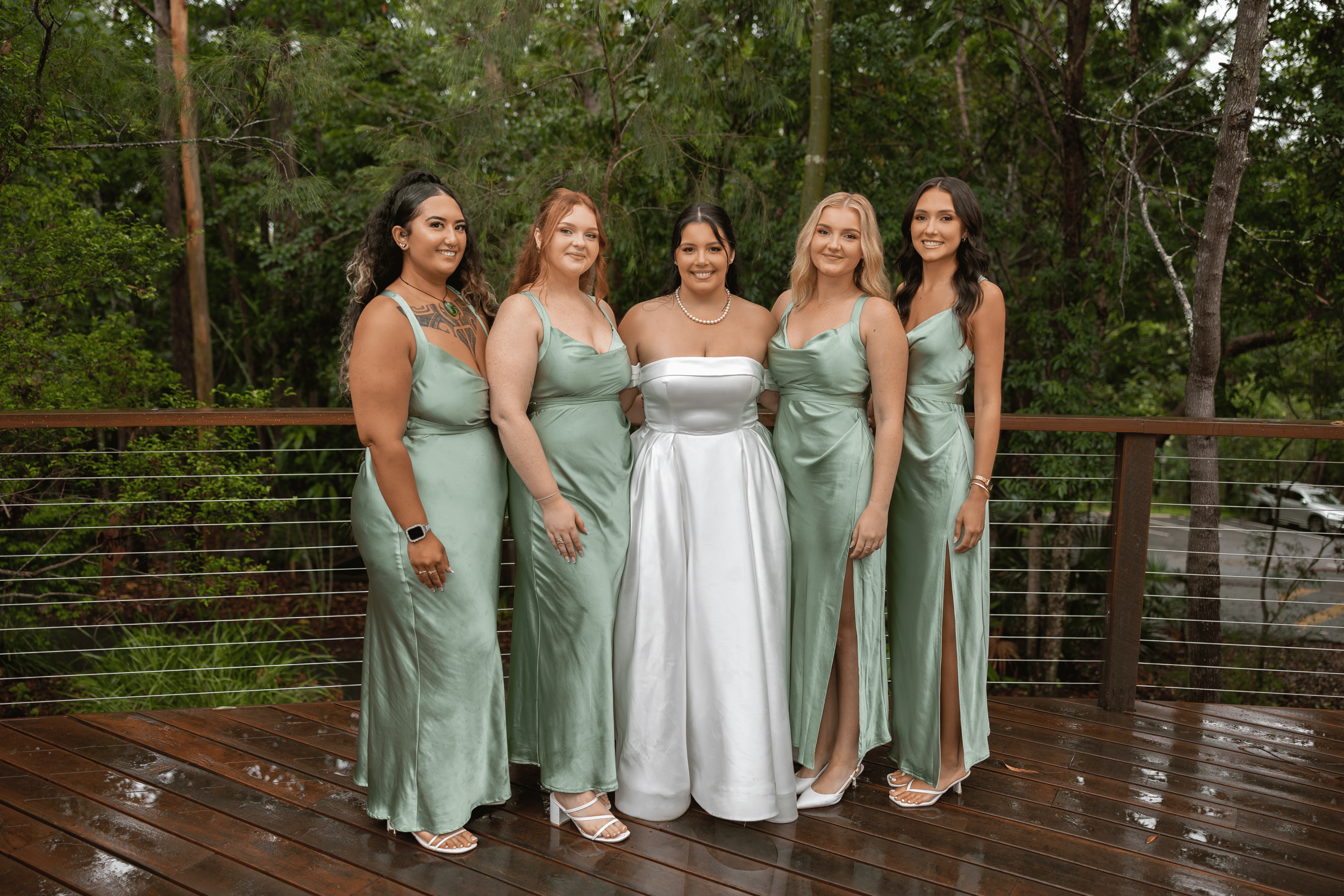 A group of six women in formal dresses standing on a wet wooden deck outdoors, with trees and greenery in the background.