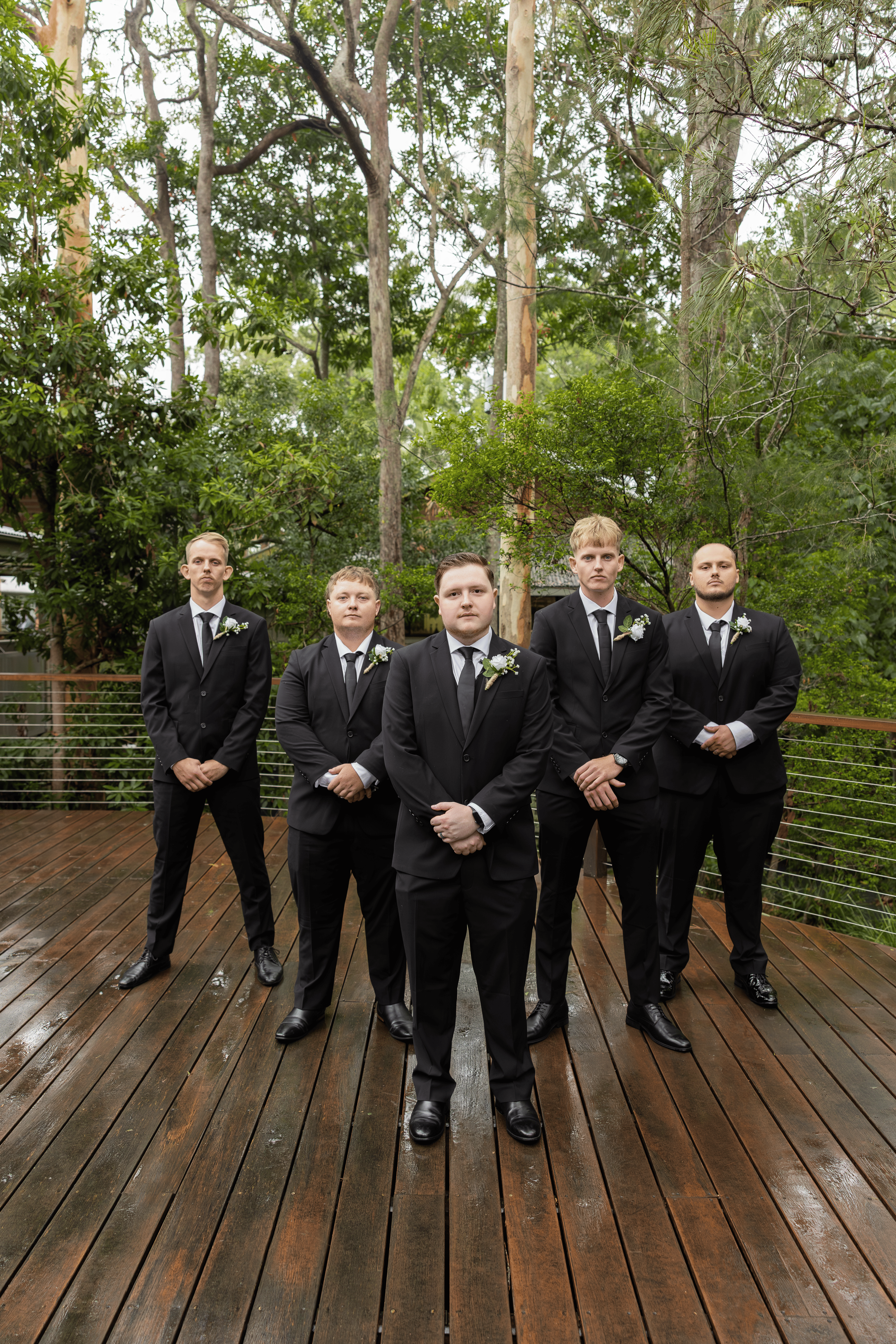 A group of five men dressed in black suits with ties, standing outdoors on a wet wooden deck surrounded by green trees, during a wedding event.