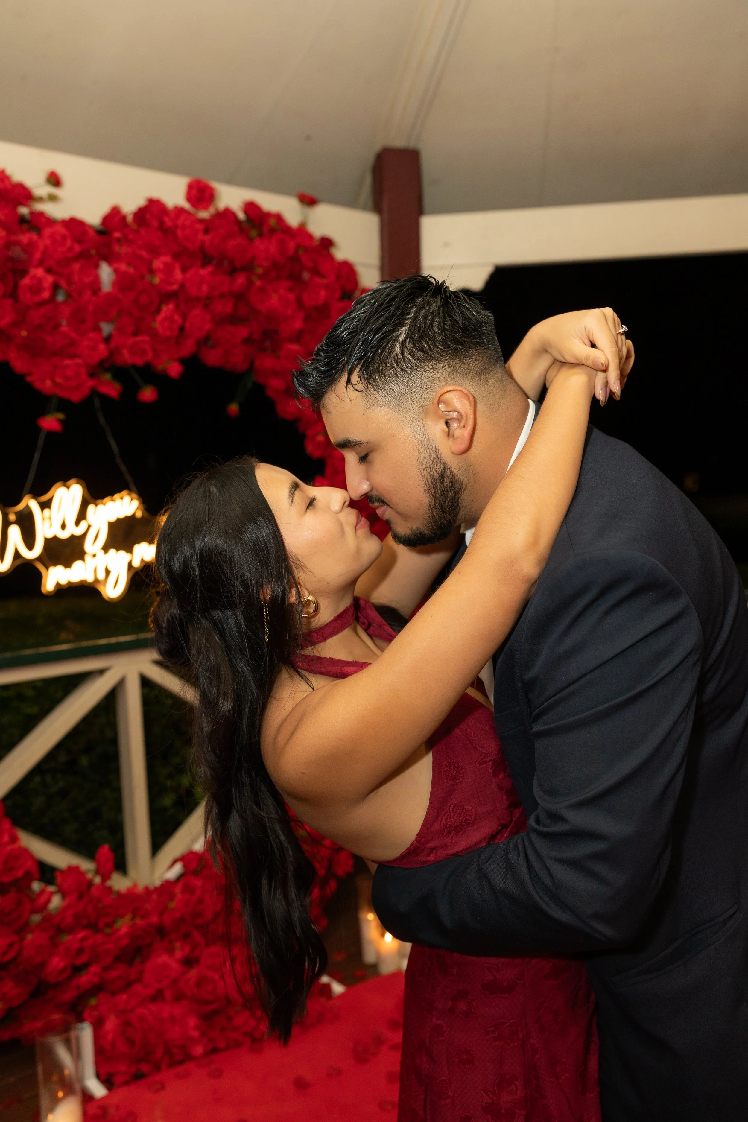 A couple dancing closely at a romantic event, surrounded by red roses and pink flowers, with a neon sign in the background.