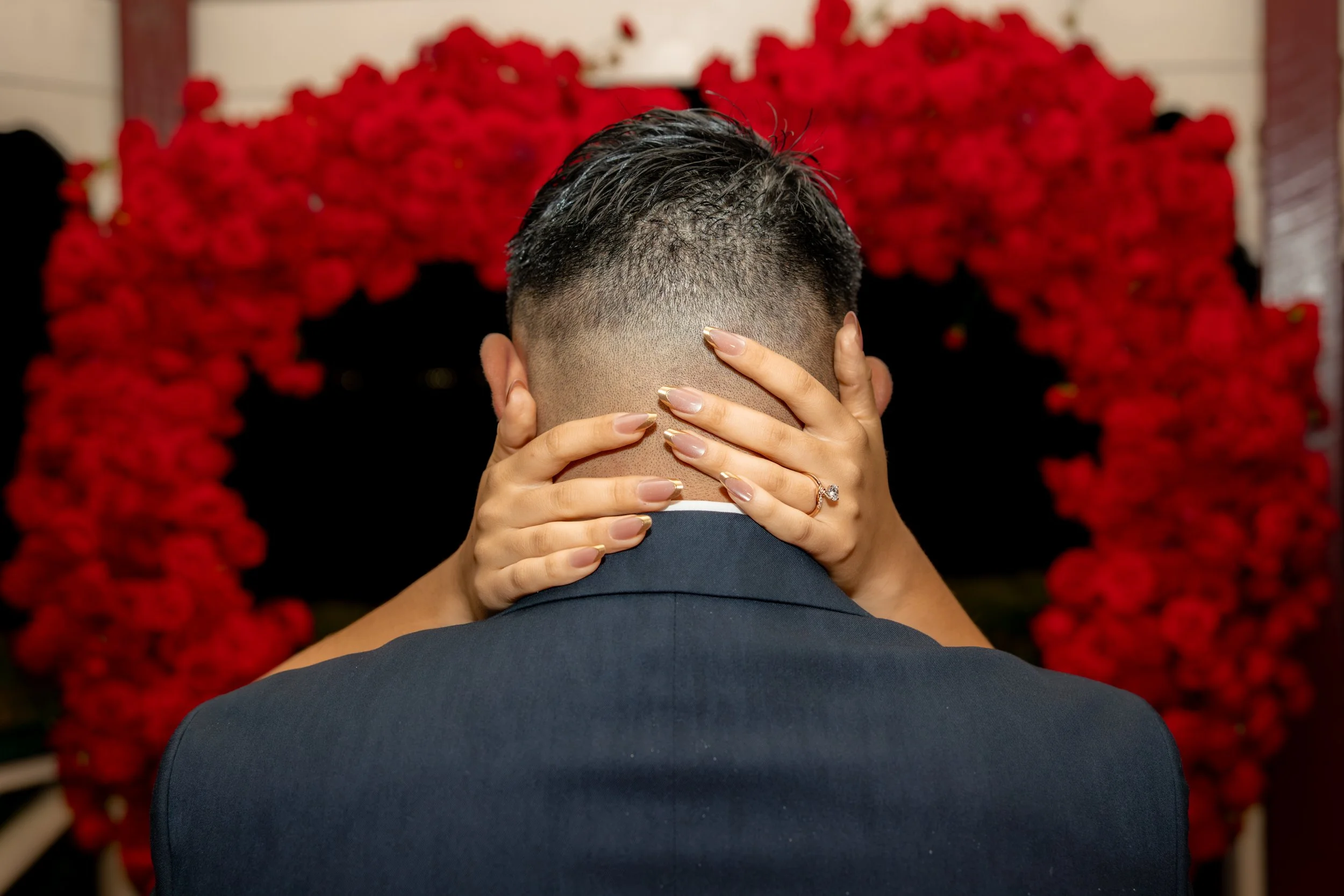 A couple embracing at a wedding, with the bride's hands on the back of the groom's neck, showing a wedding ring, in front of a red floral heart backdrop.