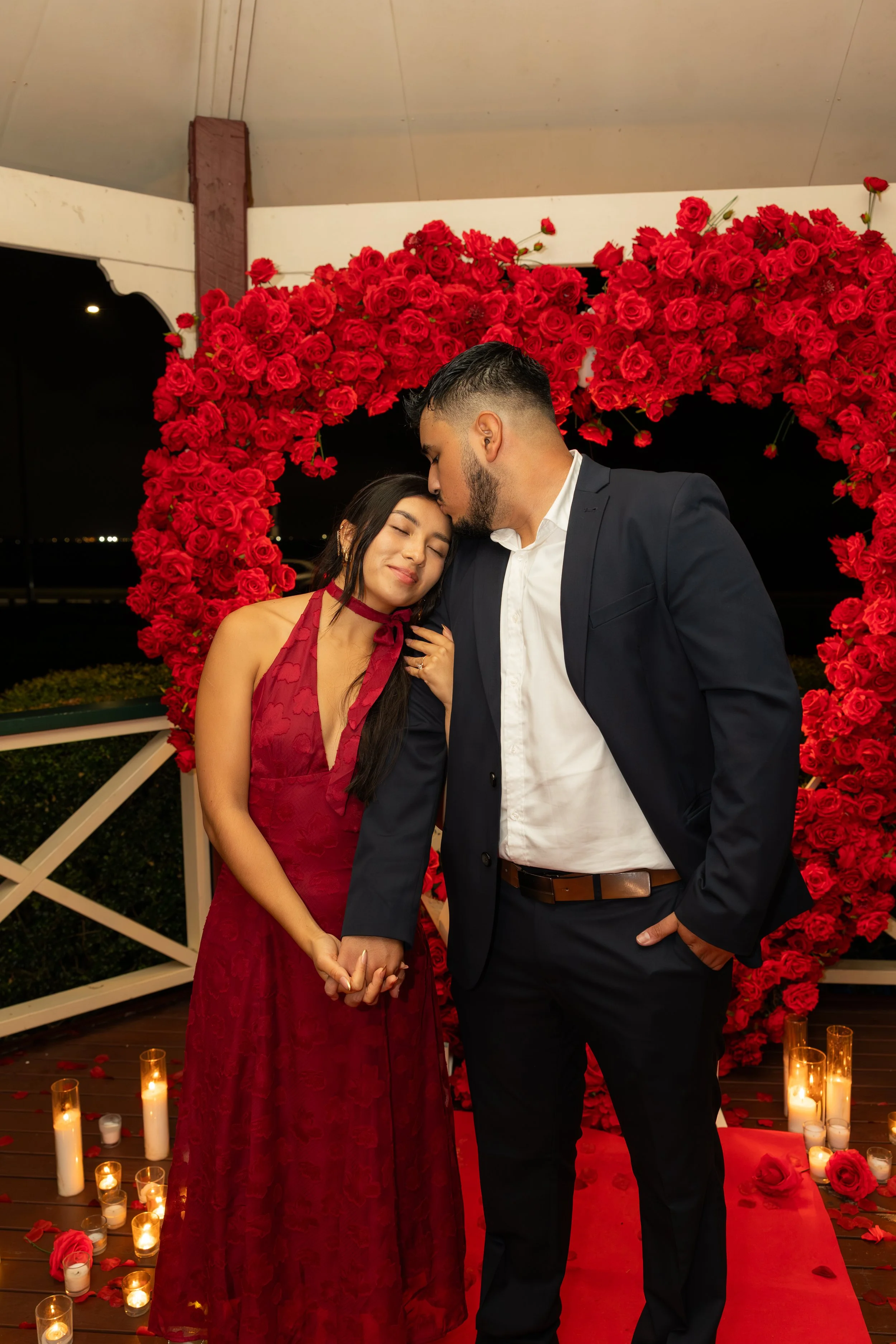 A couple holding hands, with the man kissing the woman's forehead, standing in front of a large heart-shaped arrangement of red roses, surrounded by candles on the floor, during a romantic evening setting.