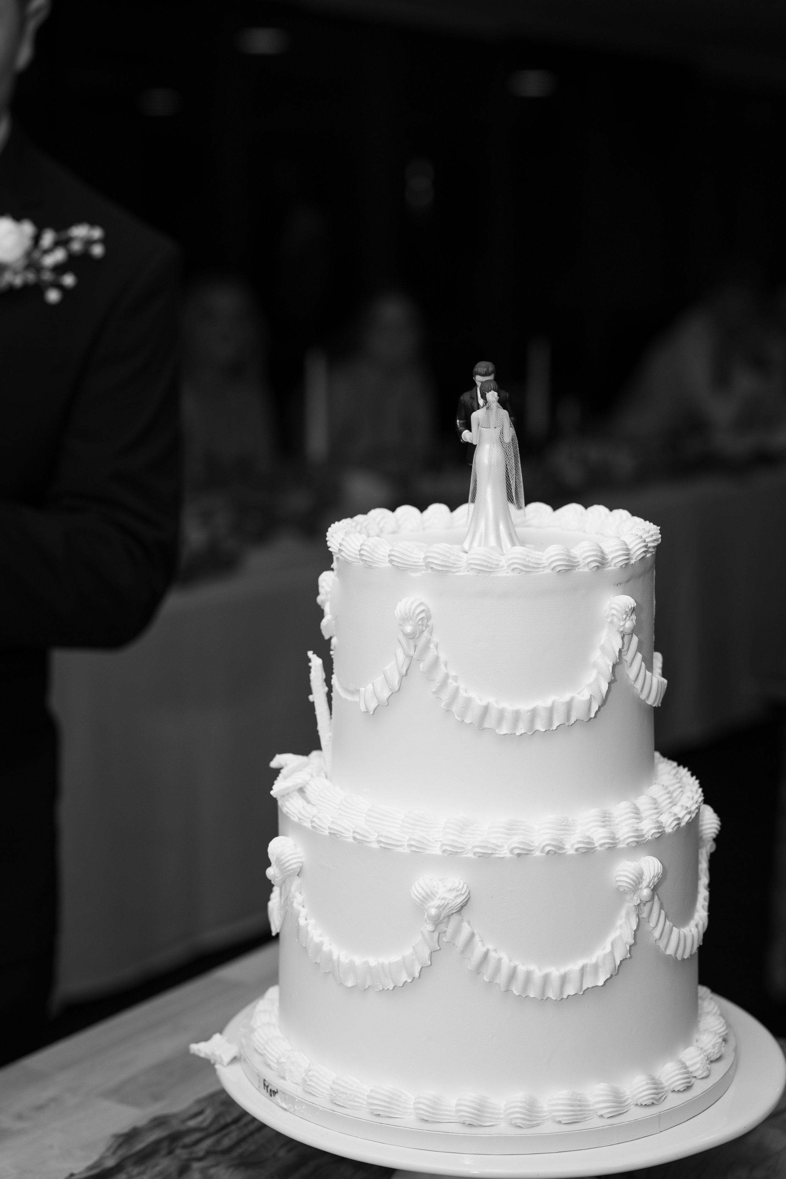 A three-tiered wedding cake decorated with white icing and garlands, topped with a bride and groom figurine.