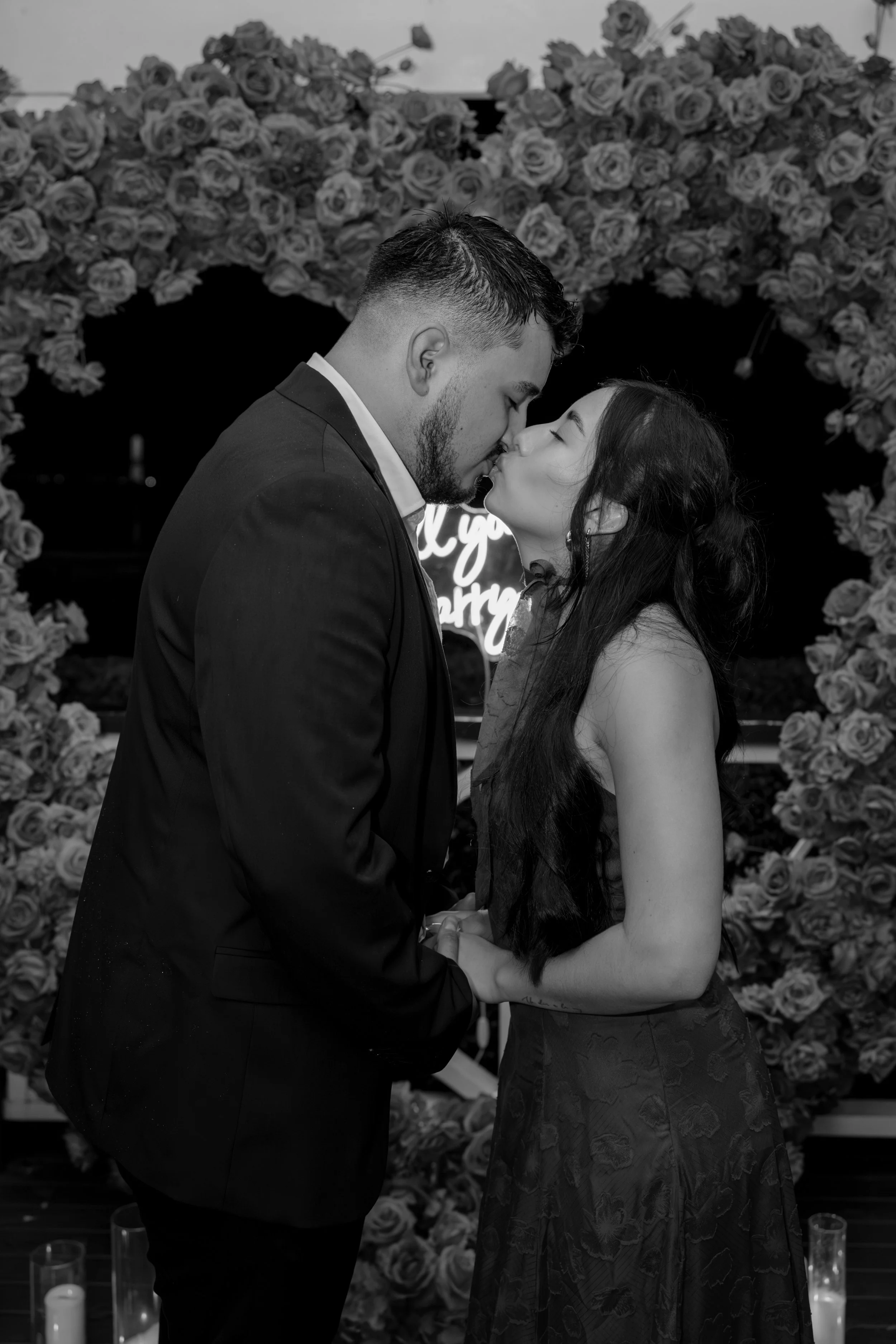 A couple in formal attire sharing a kiss at a romantic event, with floral decorations in the background.