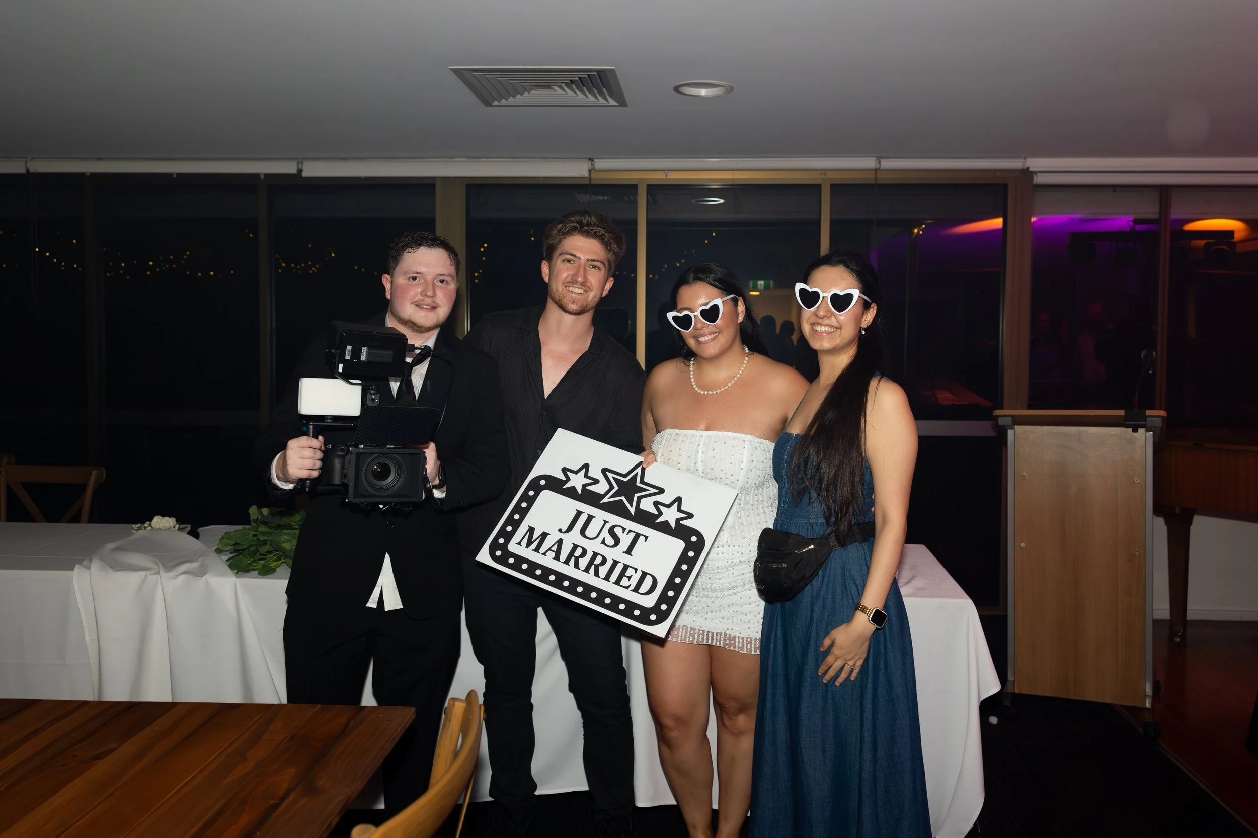 Group of four people celebrating, holding a 'Just Married' sign, at a wedding reception, with two women wearing heart-shaped sunglasses and smiling.