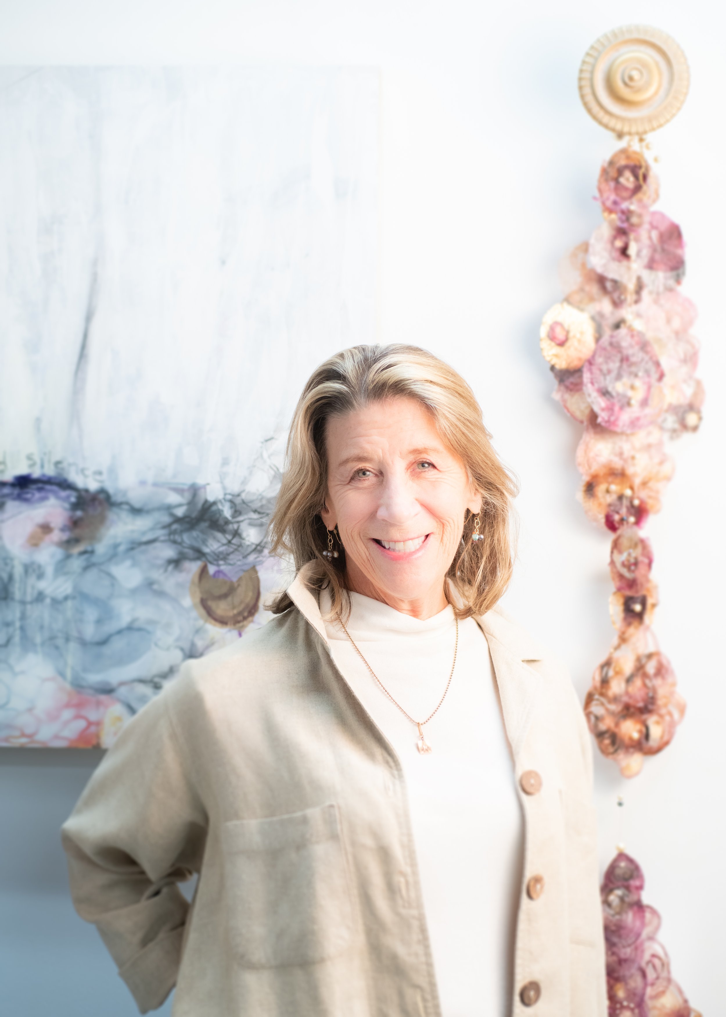 A woman with shoulder-length blonde hair and earrings smiling, wearing a beige jacket and white top, standing in front of abstract artwork and a string of large decorative seashells.