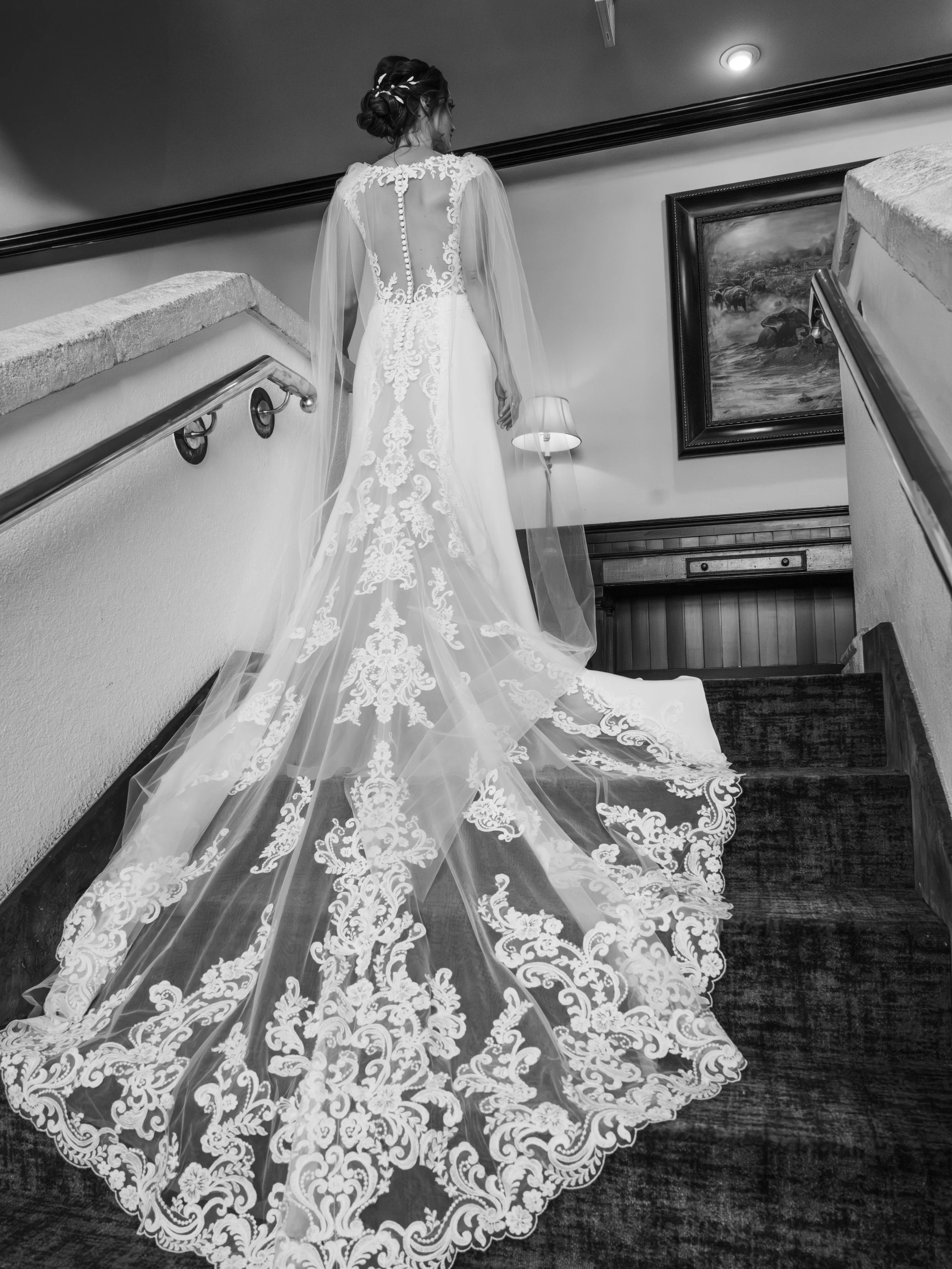 A bride in a wedding dress with a long lace train and sheer sleeves, standing on a staircase inside a house or venue.