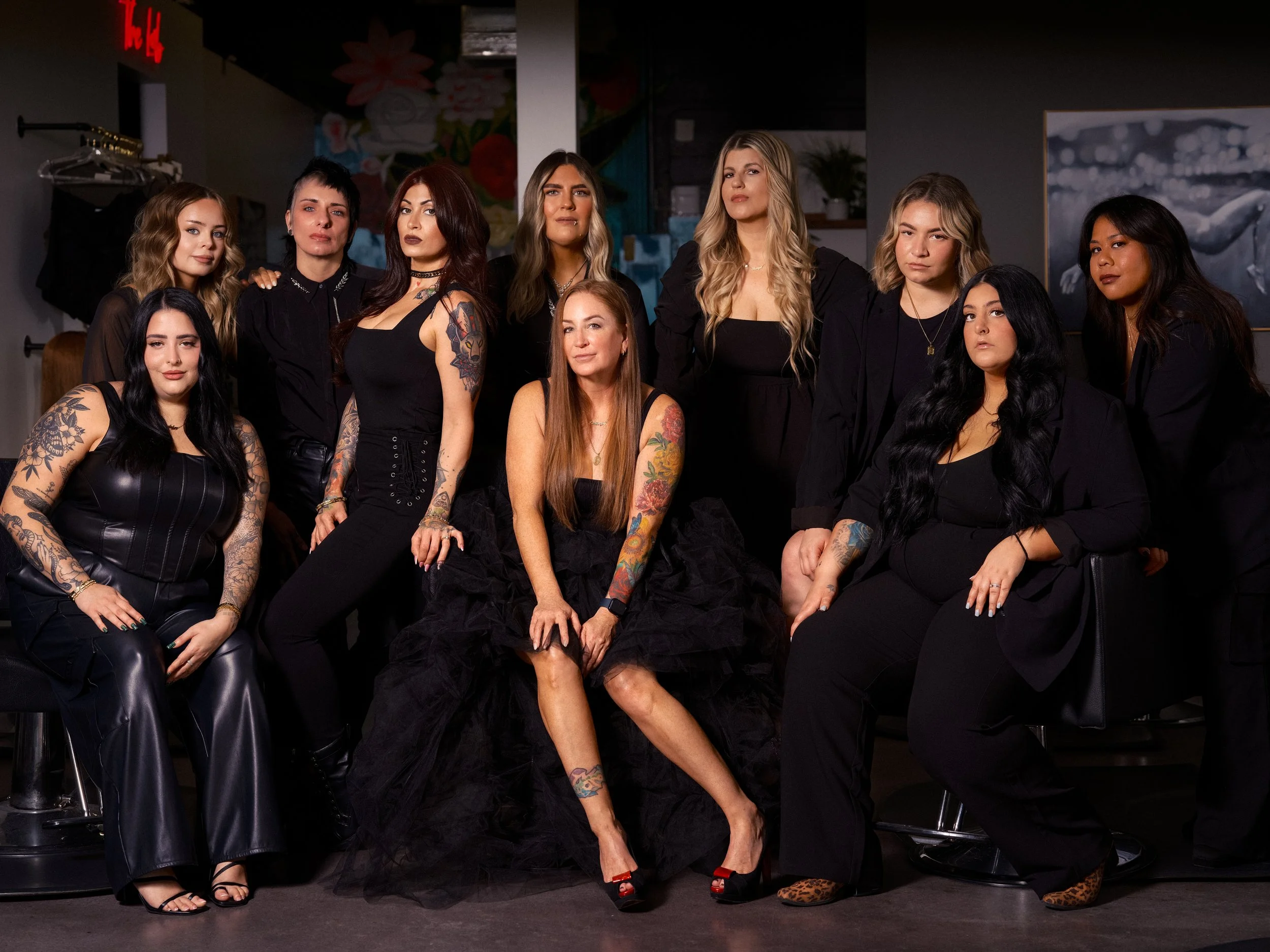 Group of eleven women with tattoos in black clothing posing together in a salon or studio