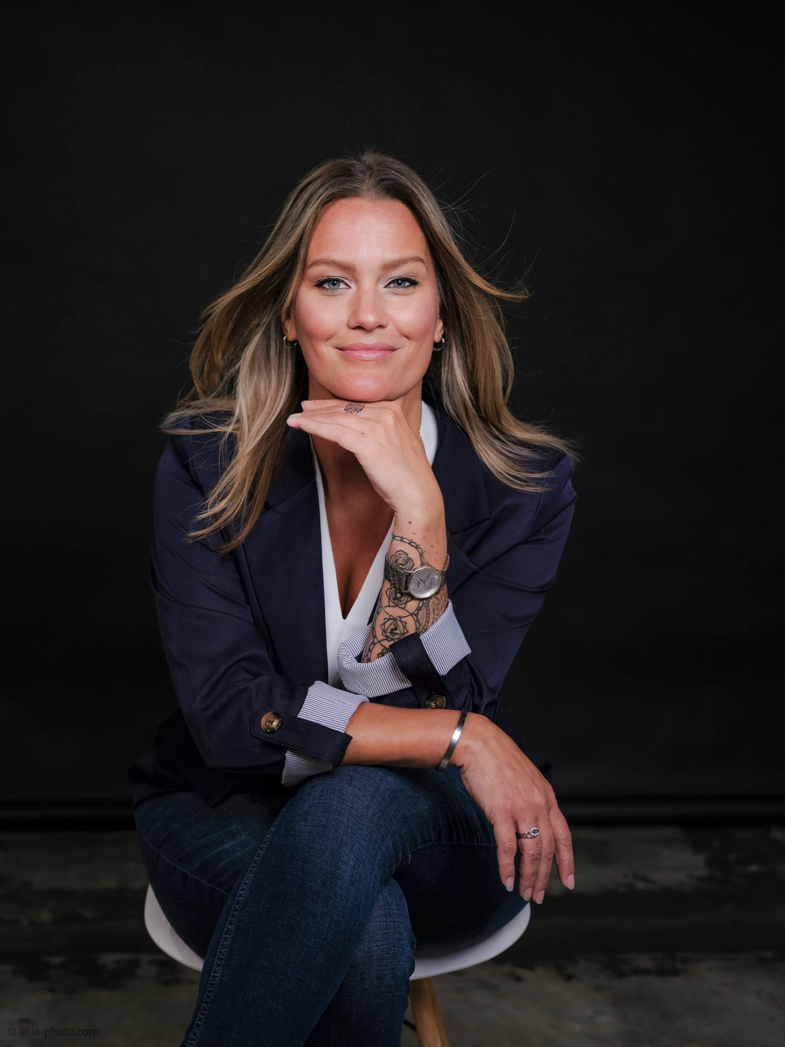 A woman with blonde hair, wearing a dark blazer and jeans, sitting on a white chair with a black background, smiling and resting her chin on her hand.