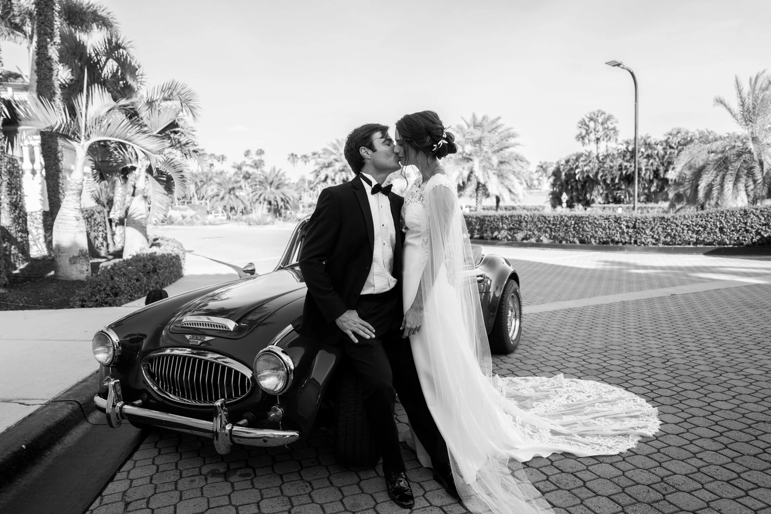 A bride and groom sharing a kiss in front of a vintage car, outdoors with palm trees and a clear sky.