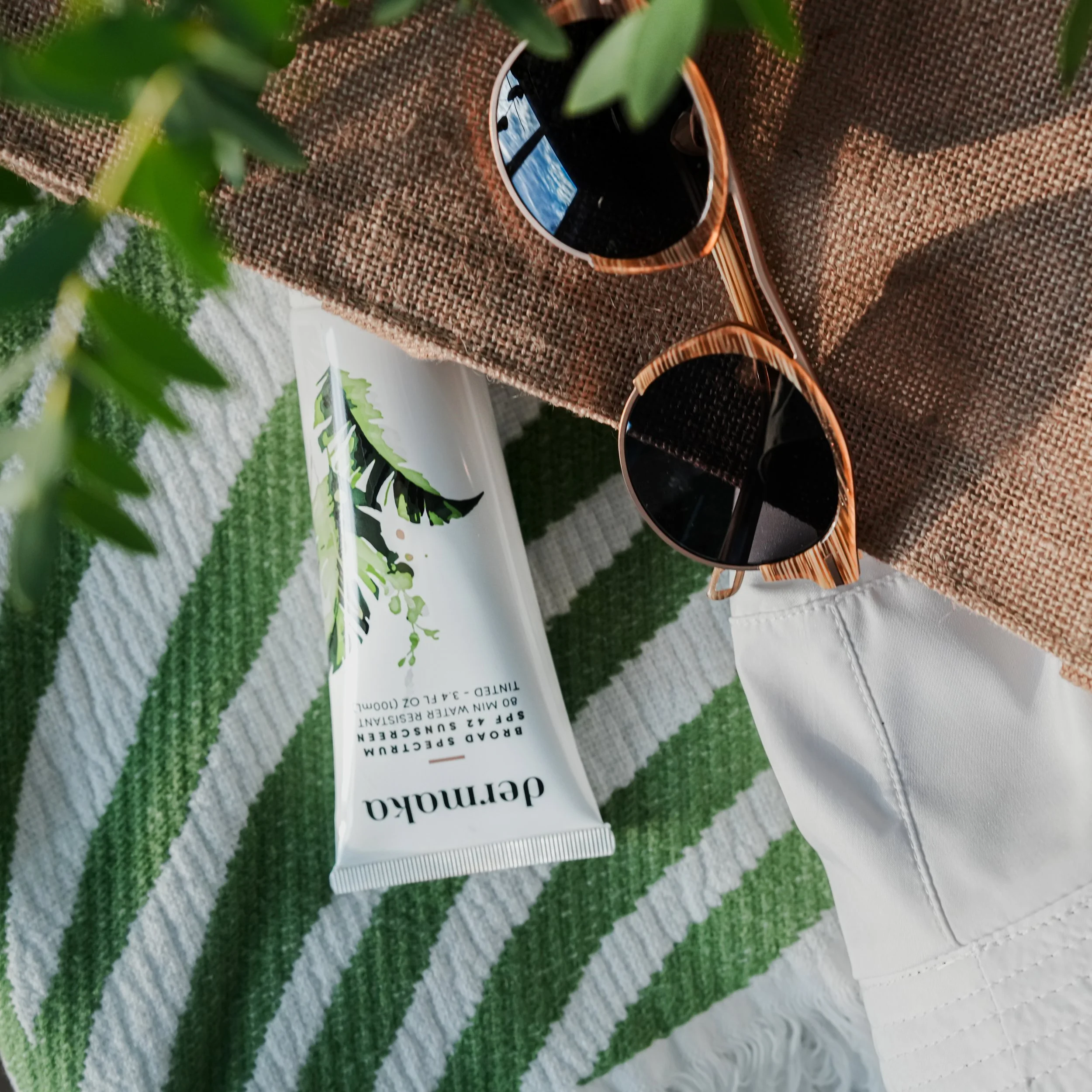 A pair of sunglasses, a tube of dermalogica skincare, a woven purse, a green and white striped towel, and part of a white fabric surface.