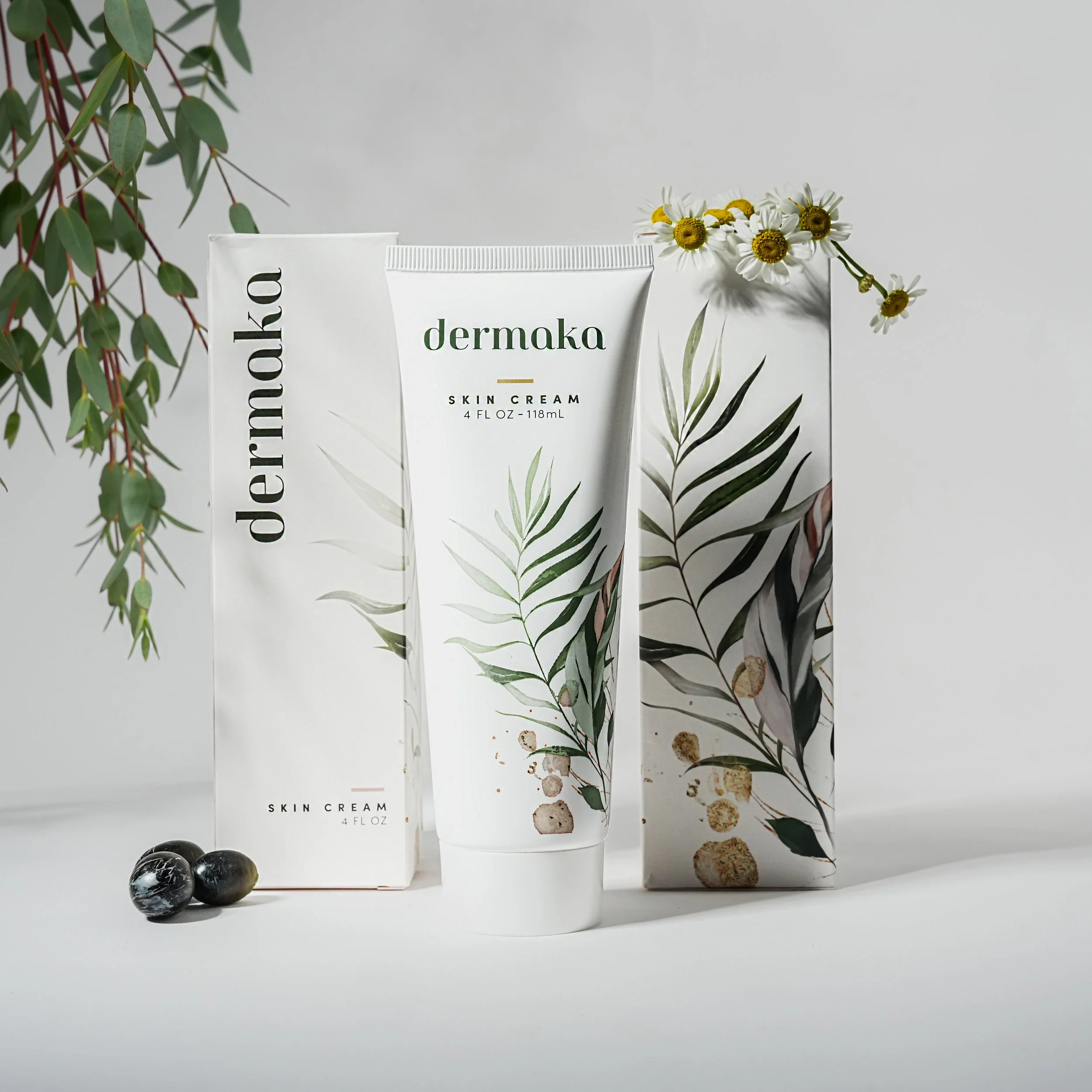 A tube of dermaka skin cream positioned in front of a matching box with botanical illustrations, accompanied by three black and white marbles and a small bouquet of white daisies with yellow centers, all against a plain and minimalistic background.