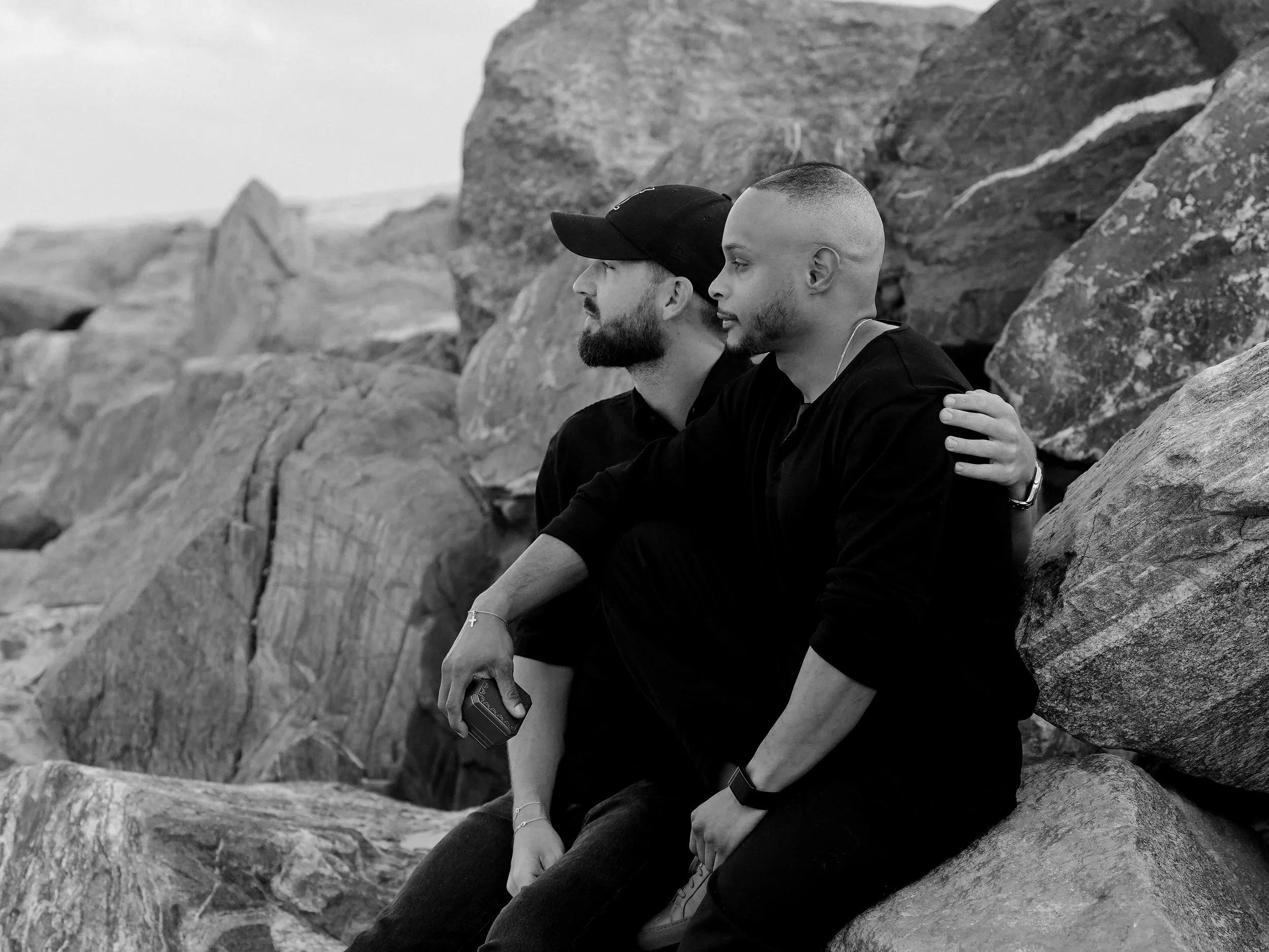 Two men sitting on rocks near a shoreline, one with a beard and cap, the other with a fade haircut, both dressed in dark clothing, one holding a phone, the scene in black and white.
